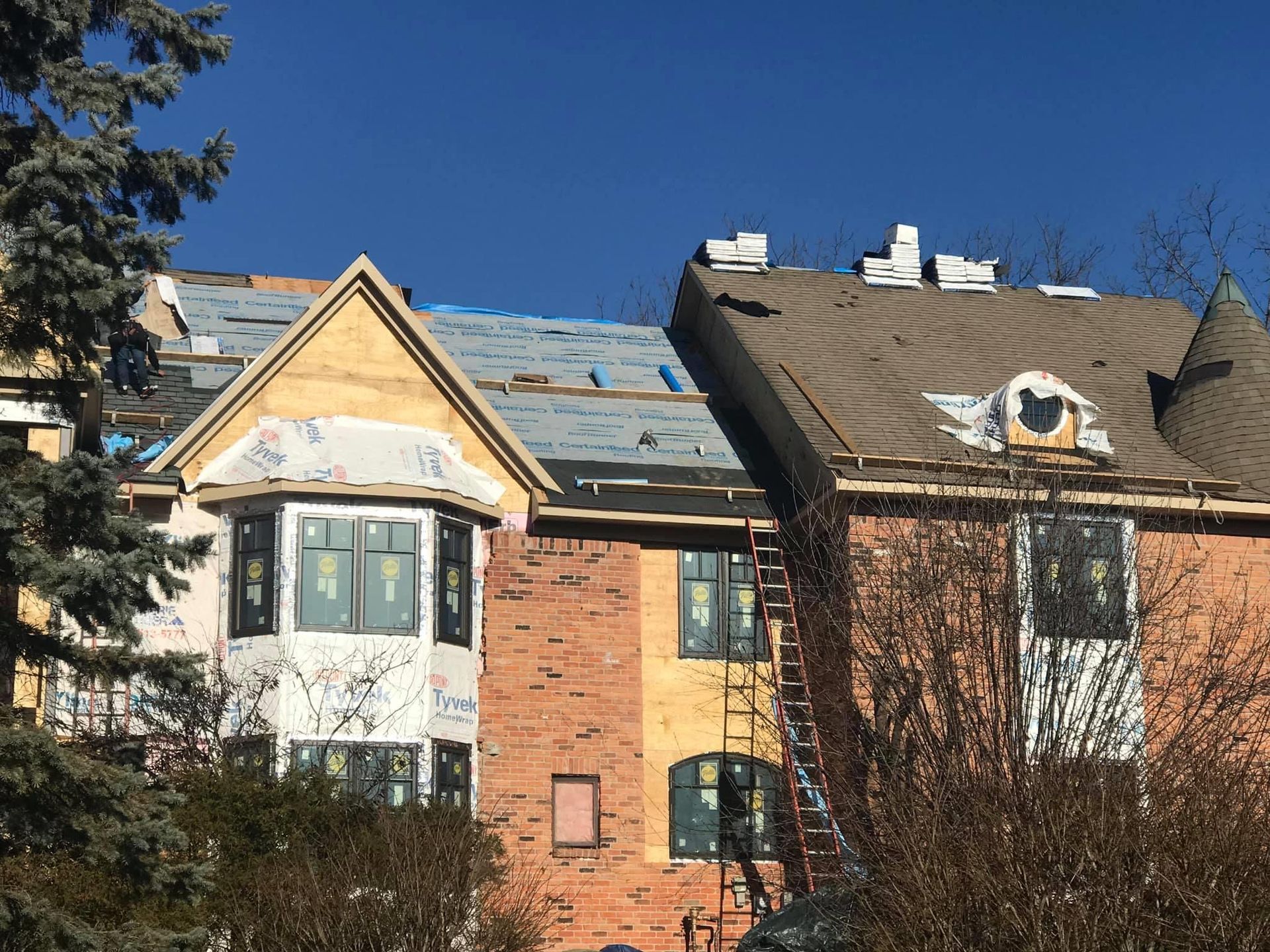 Building under construction with exposed roofing and facade, ladder, and tree against blue sky.