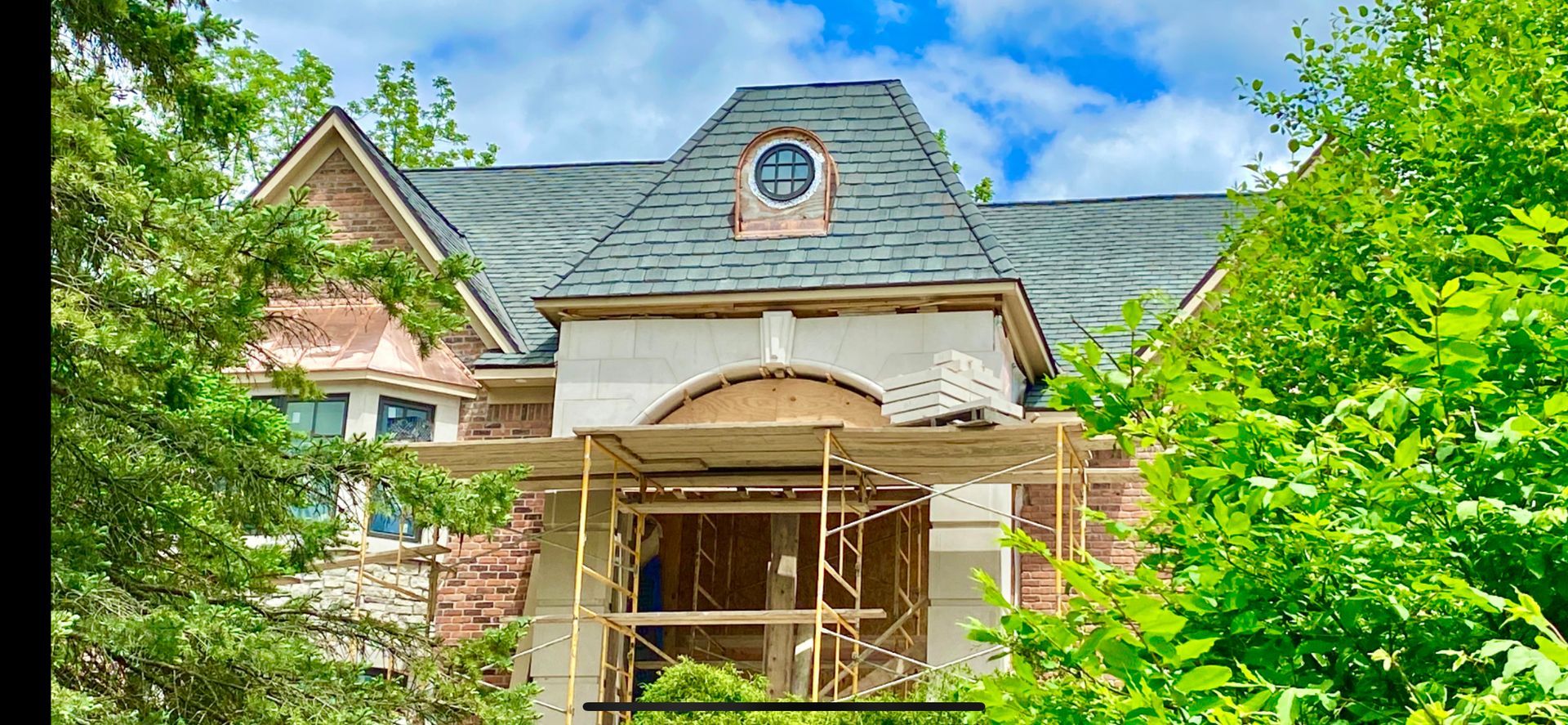 A house under construction, with scaffolding. The roof is gray and the house has a stone exterior. Green trees surround it.