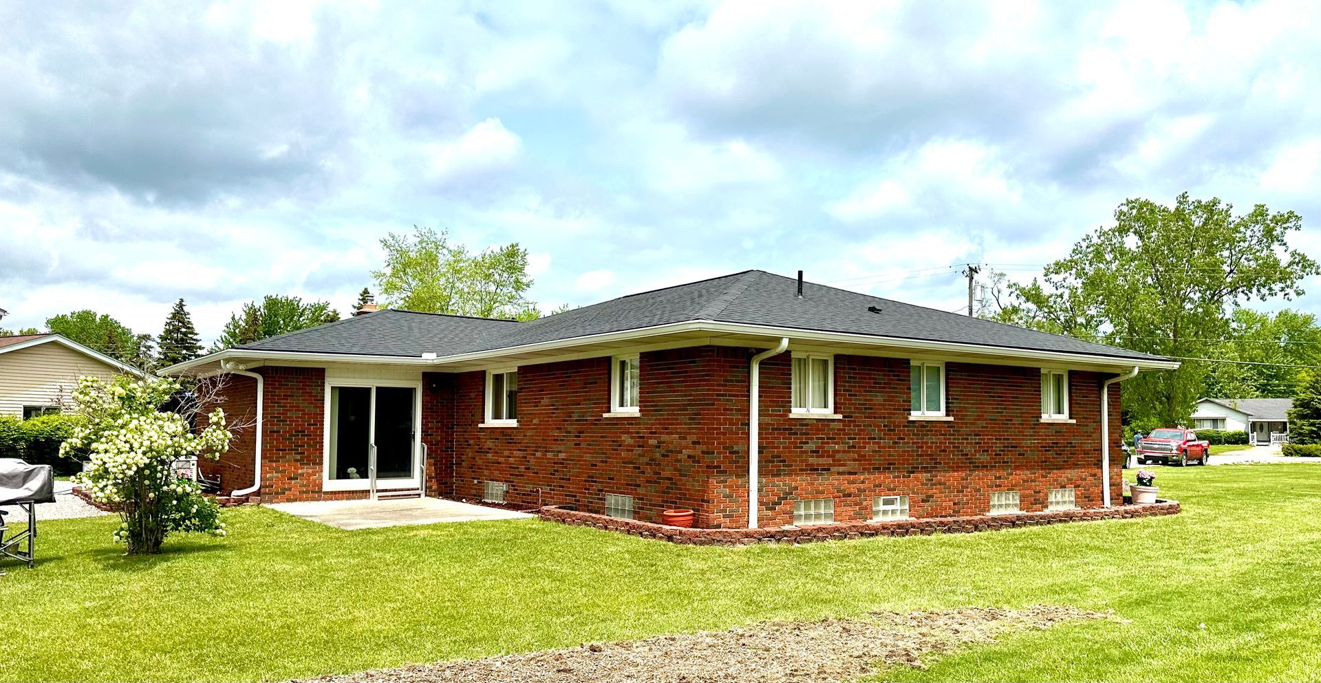 Red brick ranch-style house with dark roof and sliding glass door. Green lawn and cloudy sky.
