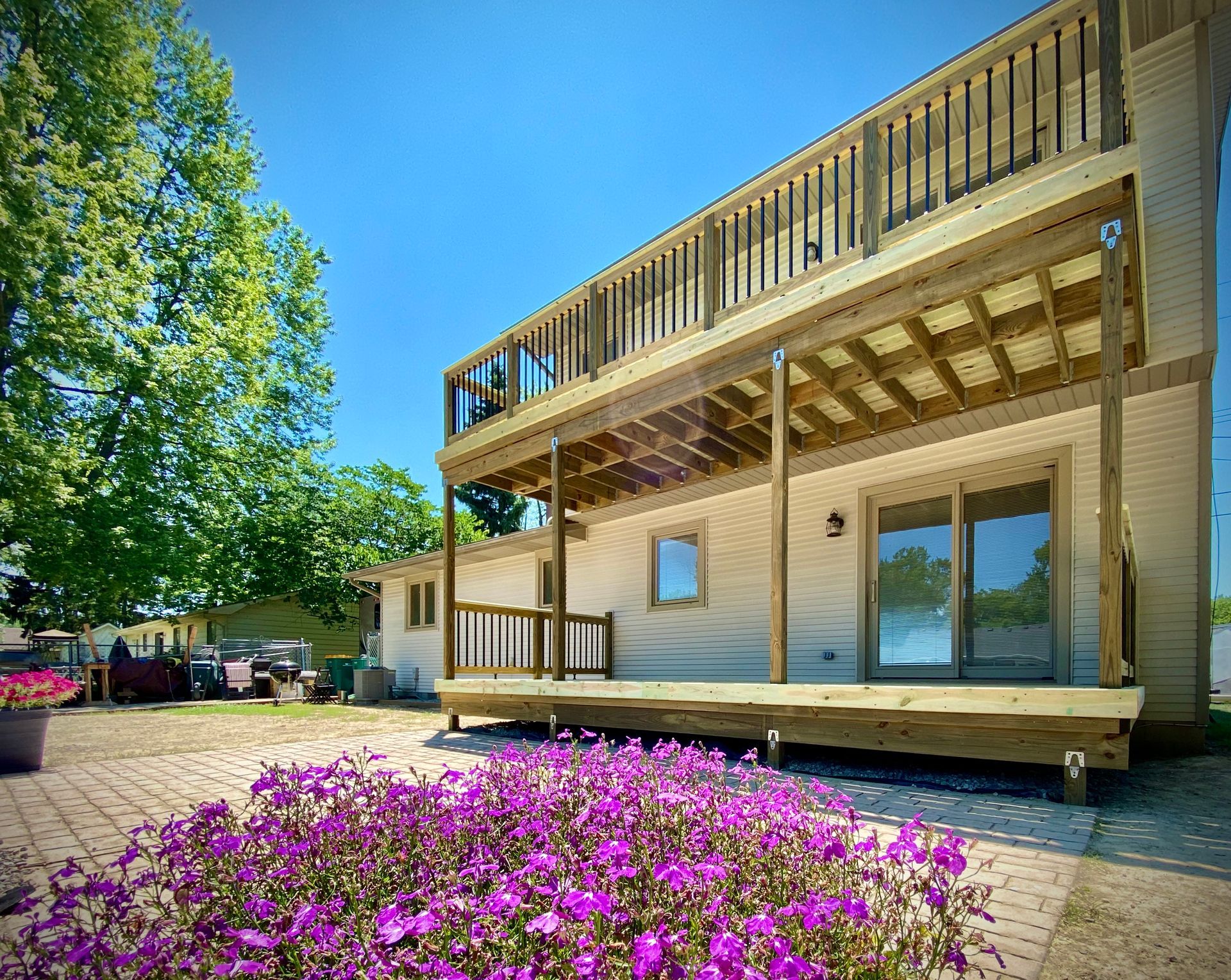 Two-story house with wooden decks. Purple flowers in foreground. Clear blue sky.