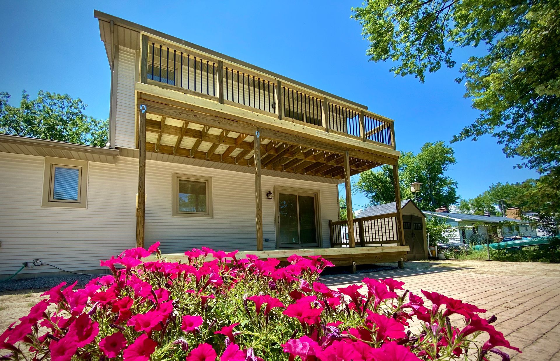 Beige house with a large wooden deck and a second-story balcony; pink flowers in foreground under a blue sky.