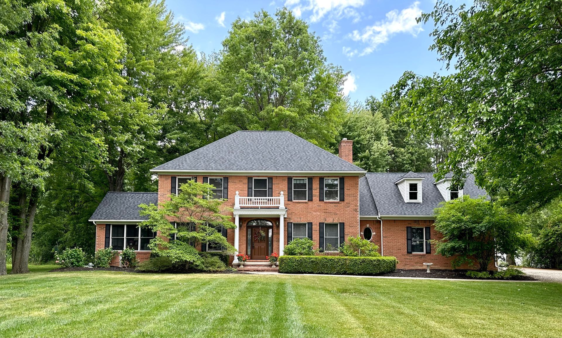 Brick house with black roof, shutters, and dormers, surrounded by green trees and lawn.