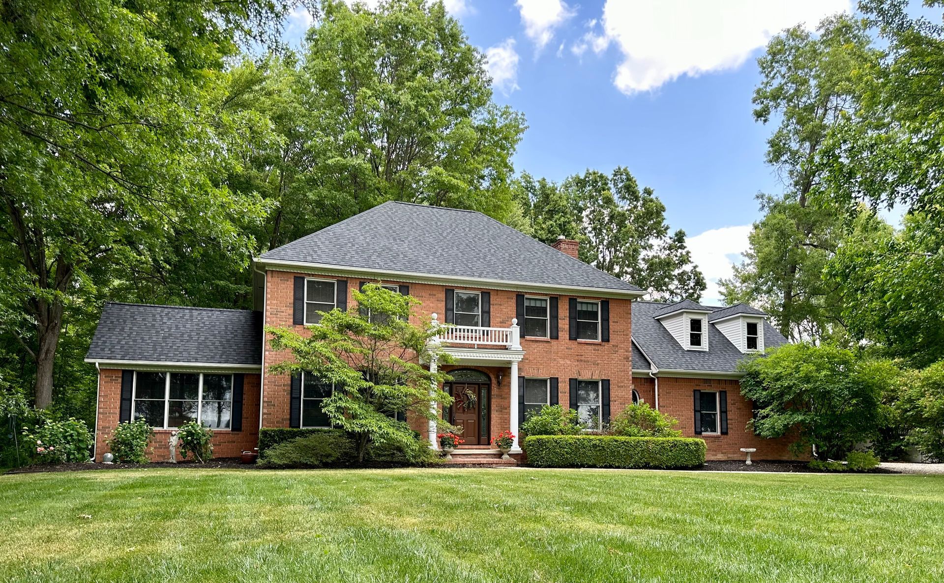Brick house with black shutters, surrounded by green trees and lawn, under a partly cloudy sky.
