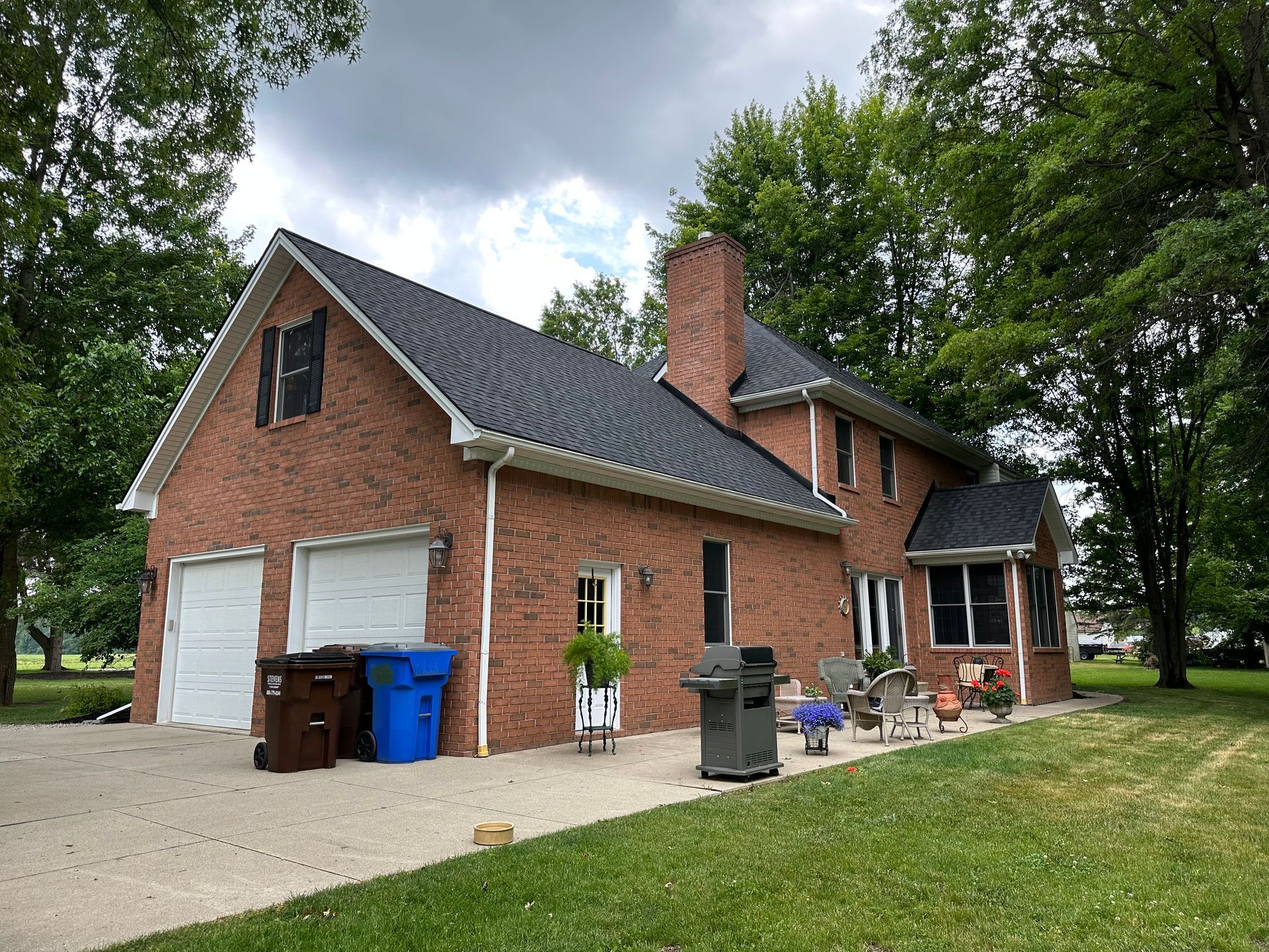Brick house with attached garage, dark roof, chimney, and backyard patio. Overcast sky.