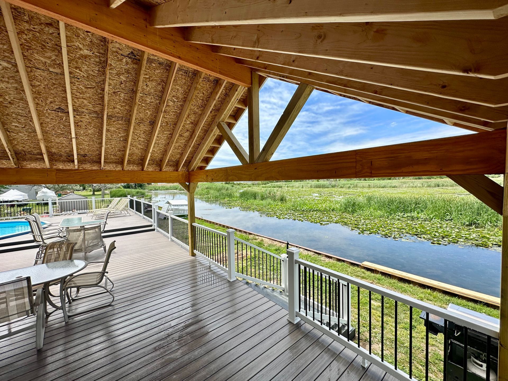 Deck with a view of a waterway, white railing, and wooden roof. Tables and chairs on the deck. Blue sky.