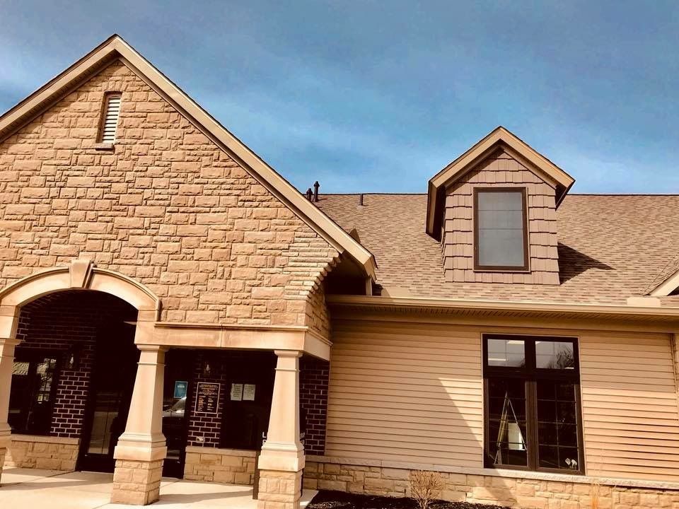 Tan stone and beige siding building with brown roof under a blue sky.