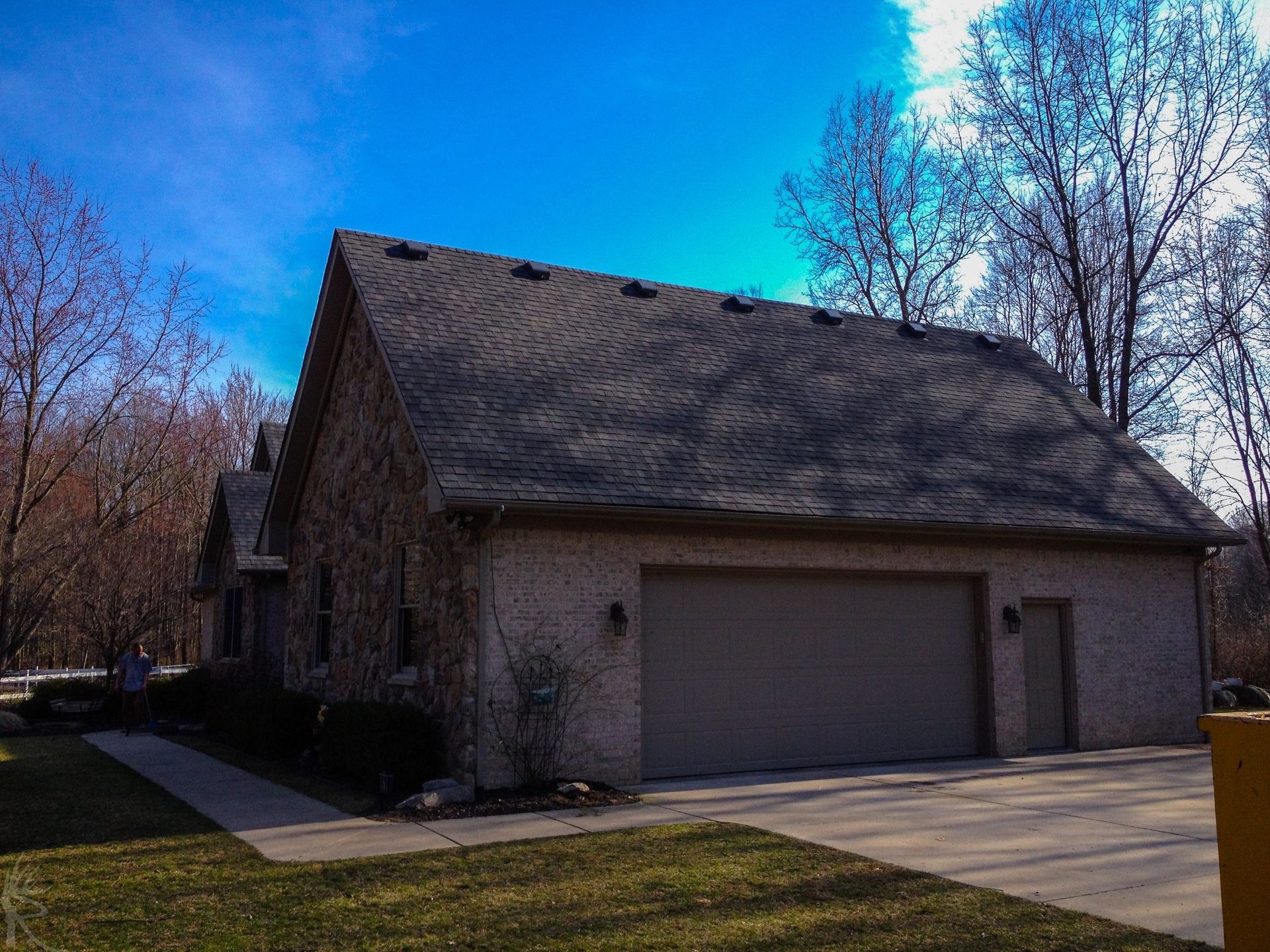 Brick house with a two-car garage under a blue sky, surrounded by bare trees and green grass.