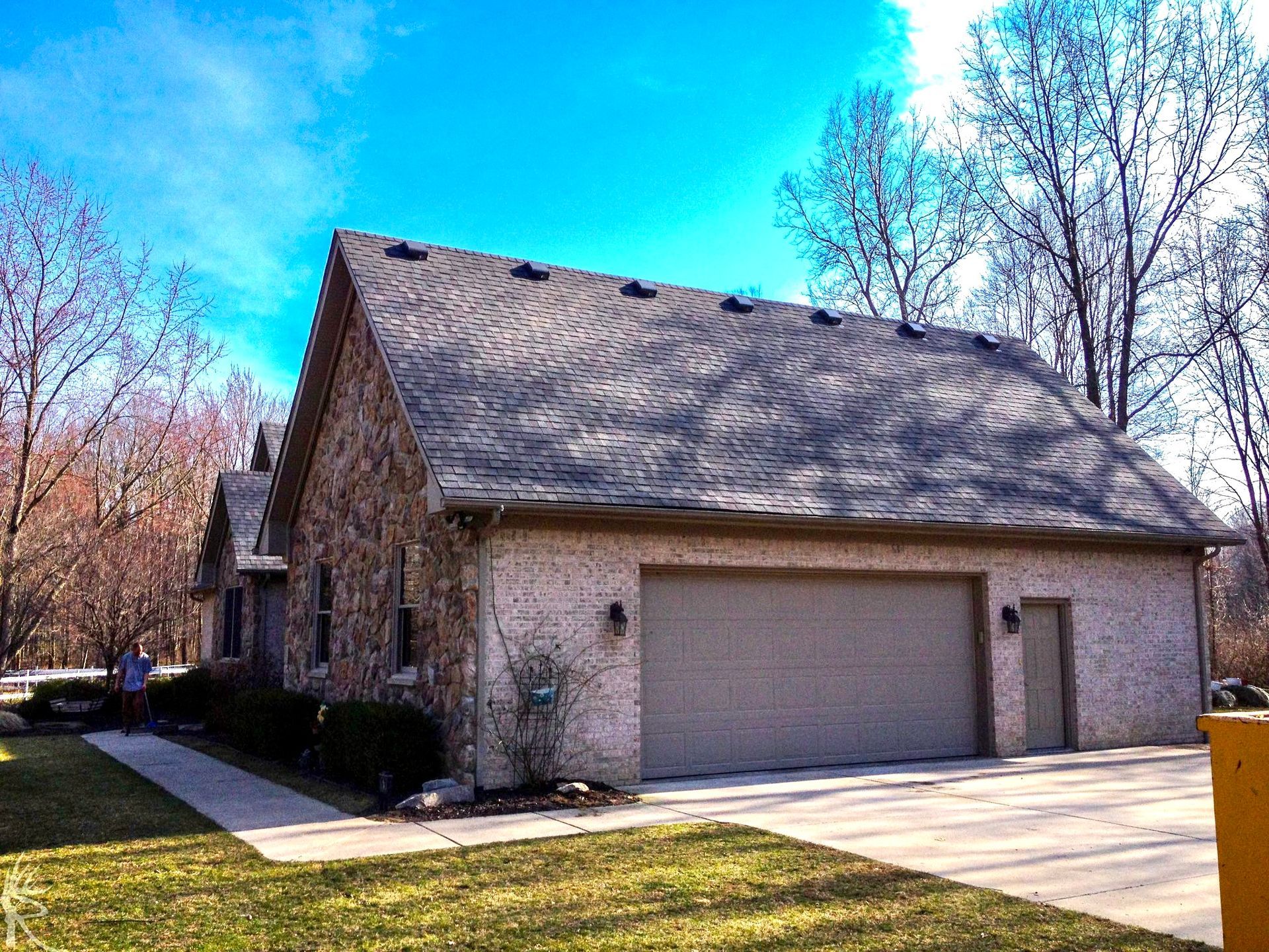 Brick house with a garage and tan door, brown roof, and trees under a blue sky.