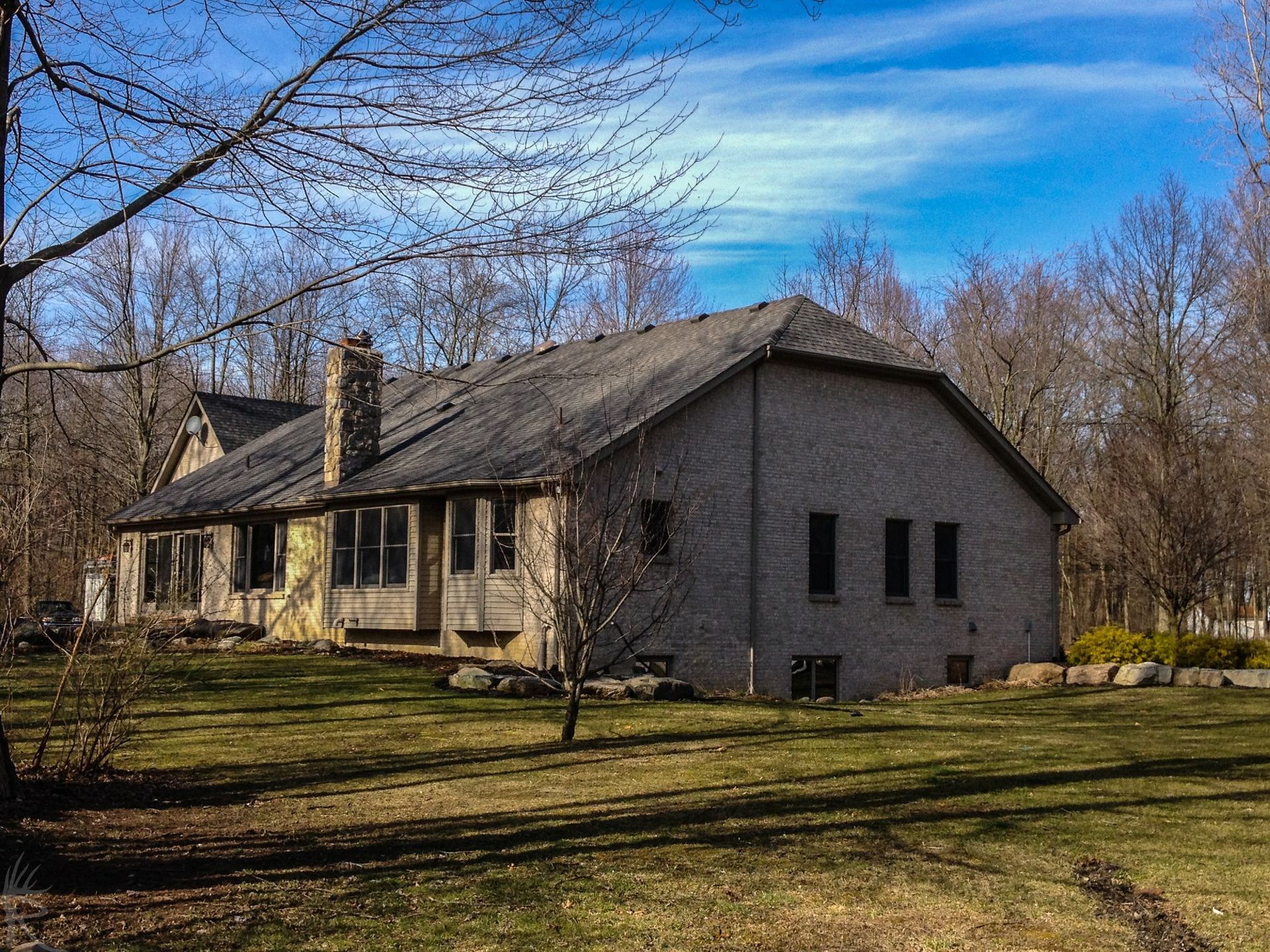 A single-story brick house with a dark roof and a chimney, surrounded by leafless trees and grass under a blue sky.
