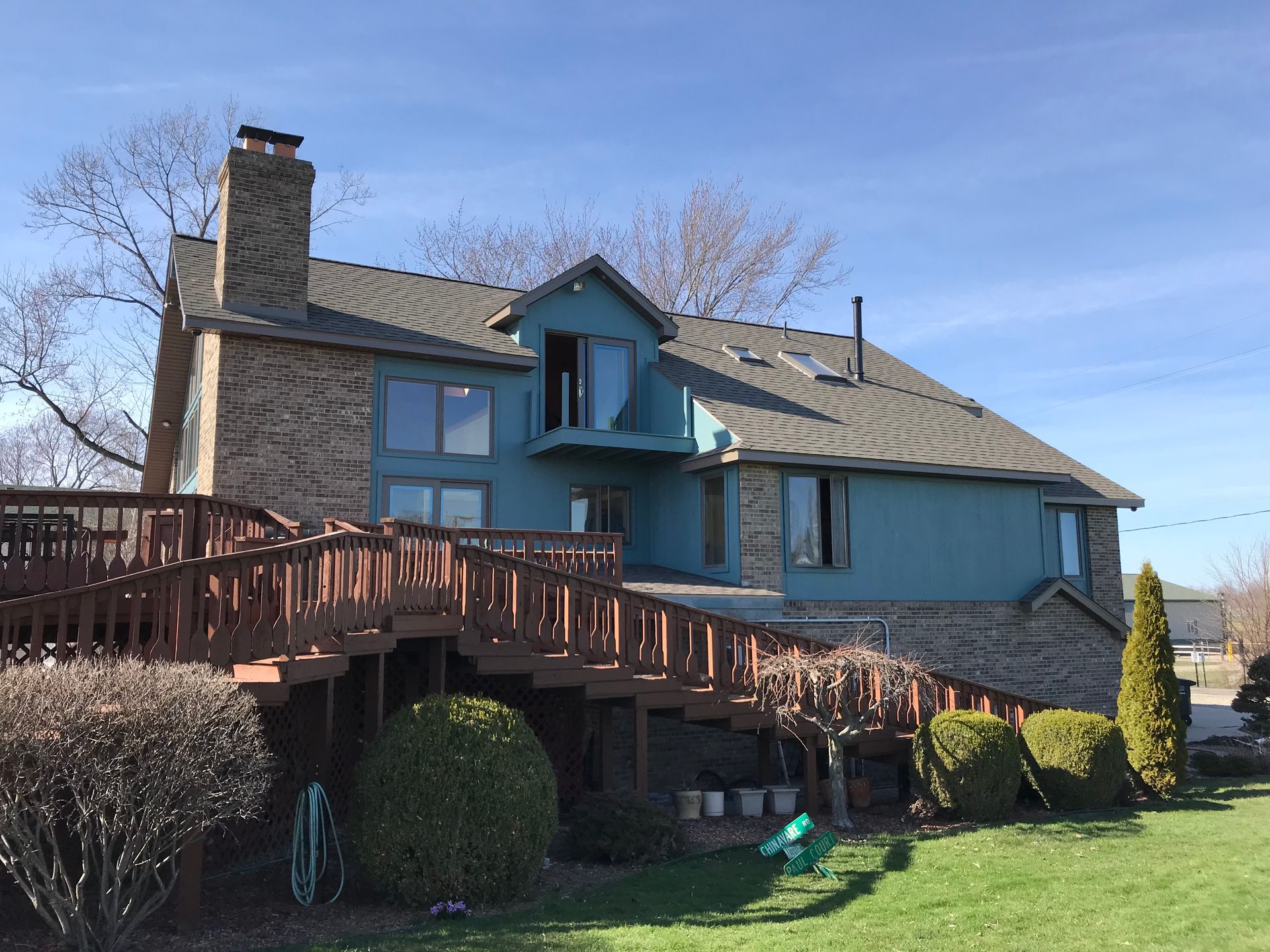 House with a blue exterior and brick, large wooden deck, green lawn, and chimney against a blue sky.