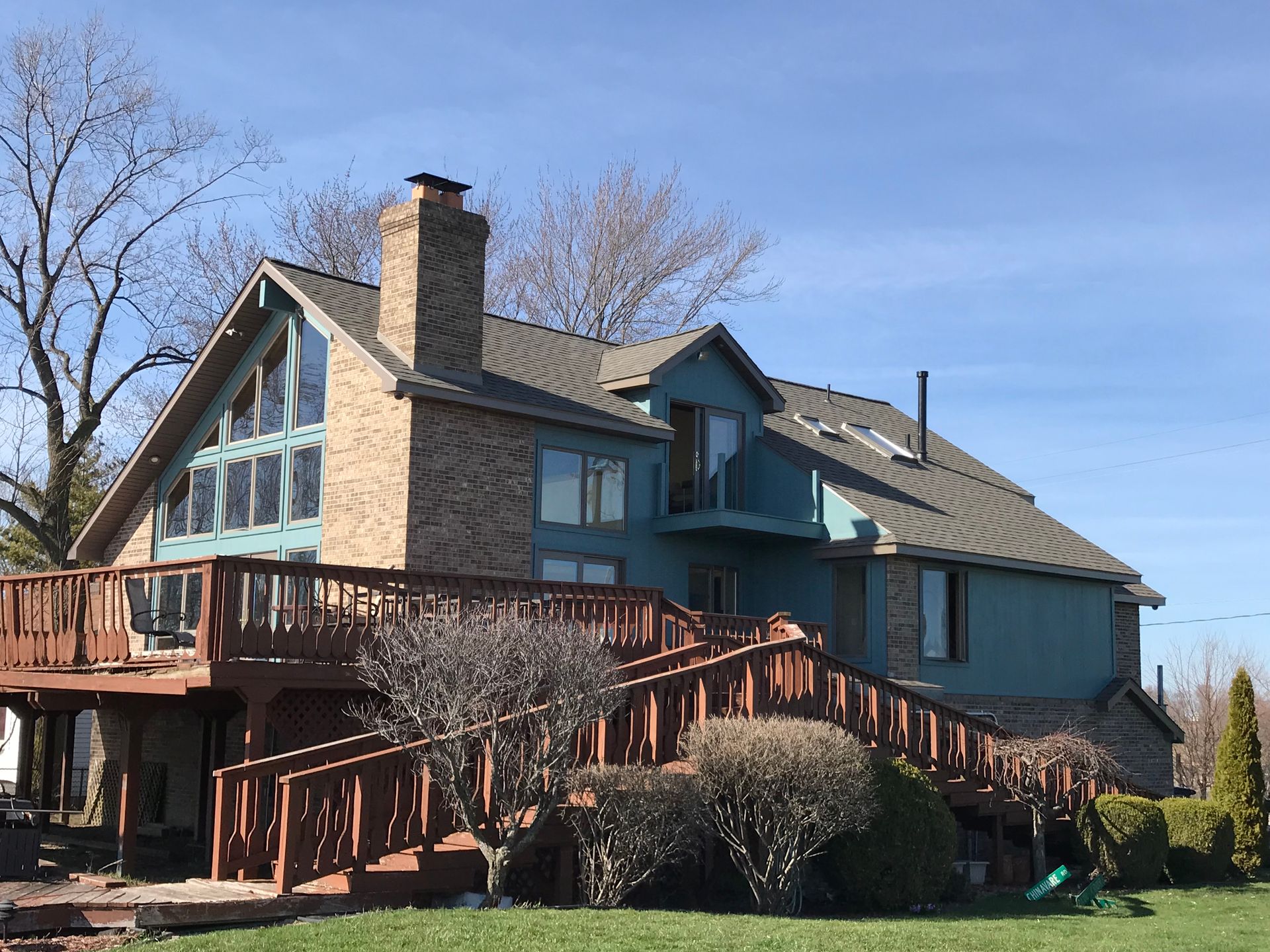 A two-story teal house with a large wooden deck and a brick chimney under a blue sky.
