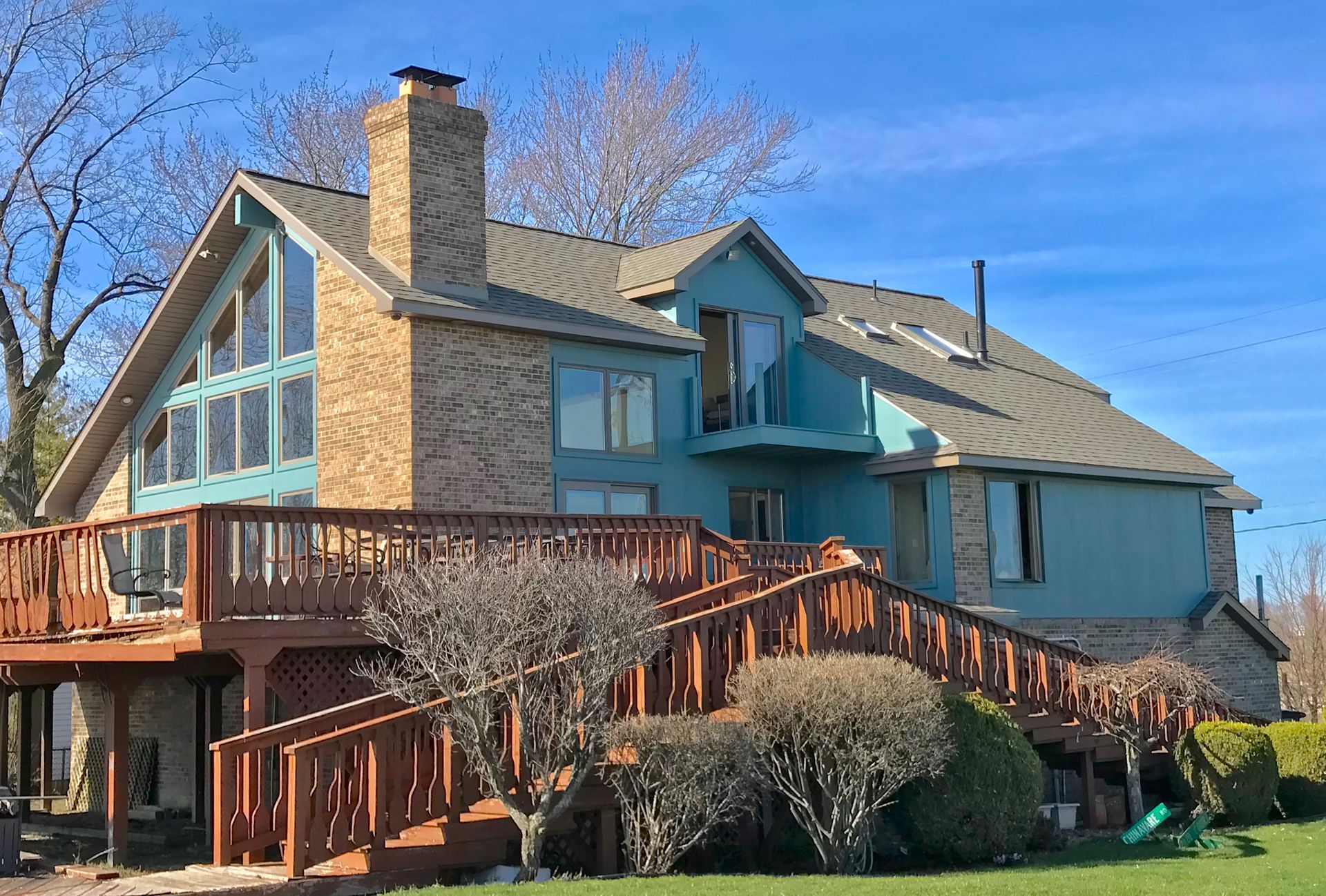 House with wooden deck and teal siding, stone chimney, and large windows against a blue sky.