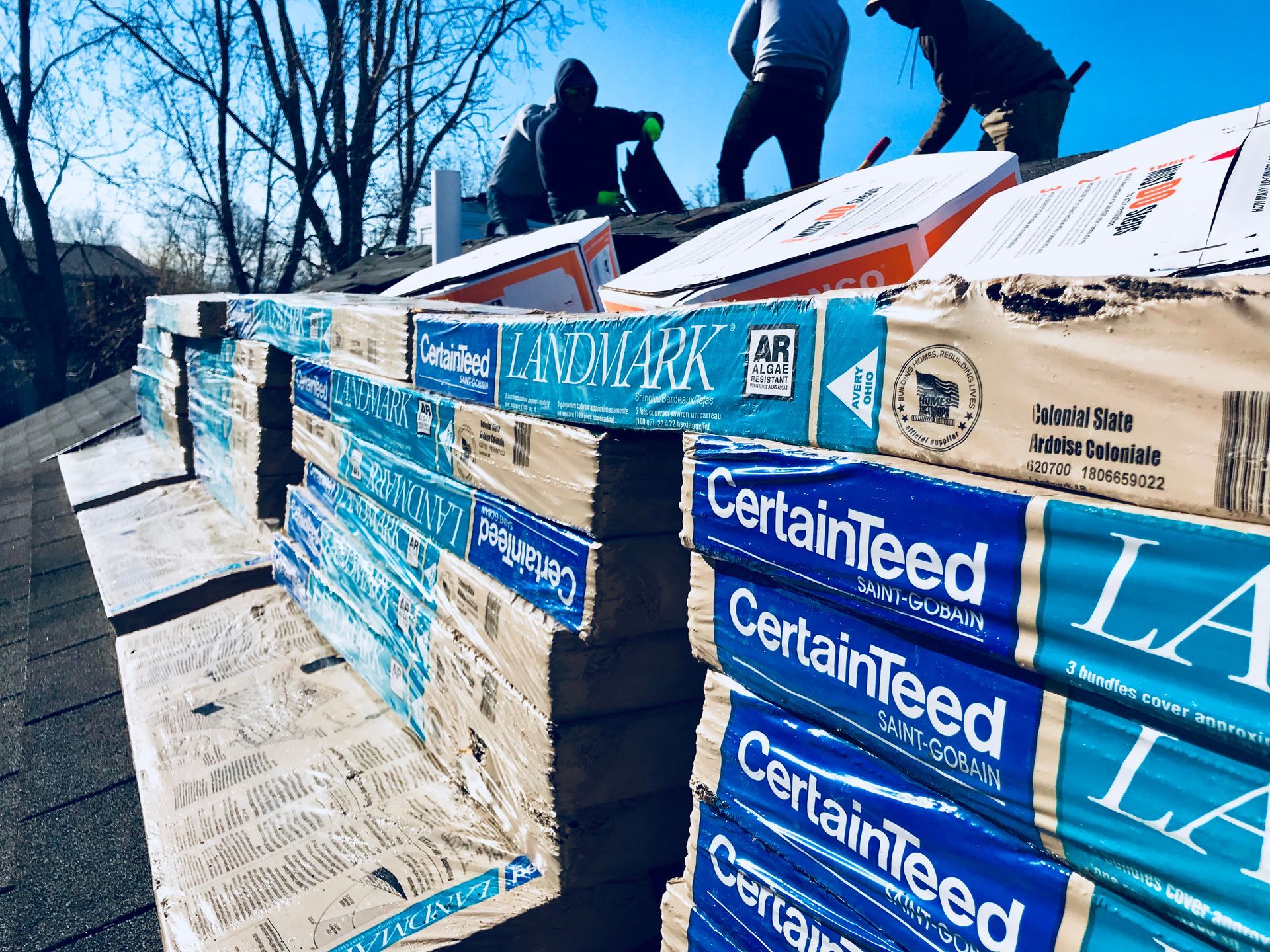 Roofers installing CertainTeed Landmark shingles on a rooftop, with stacks of materials and blue sky.