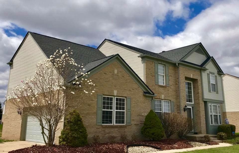 Two-story brick house with green trim, white siding, and a cloudy sky background.