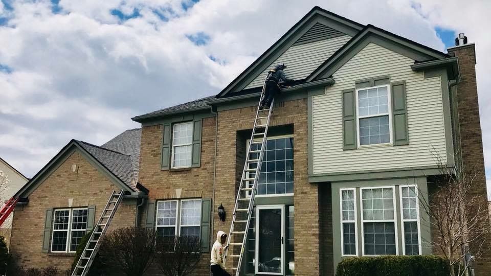 Person on tall ladder cleaning gutters of a two-story brick house with green shutters and siding.