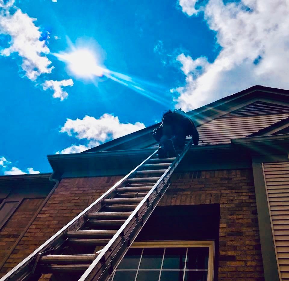 Person on ladder reaching roof of brick building under a sunny sky.