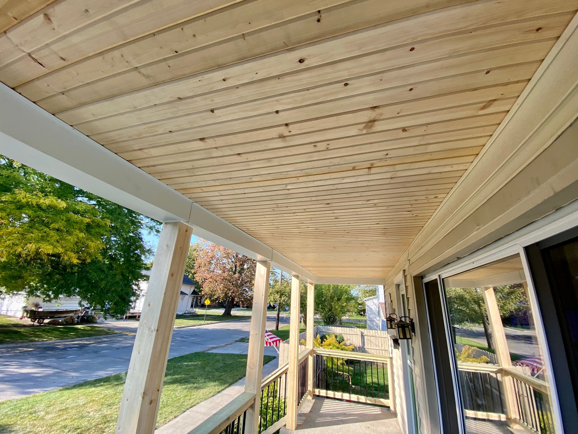 Porch with wood ceiling and white trim. View of a street with trees and homes in the background.