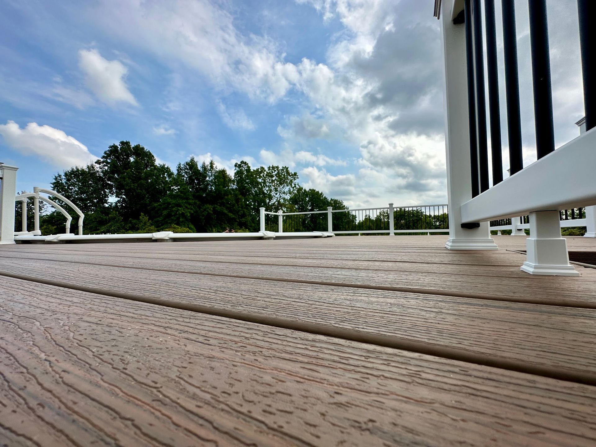 Low-angle view of a wooden deck with white railing, black vertical accents, against a cloudy sky with trees.