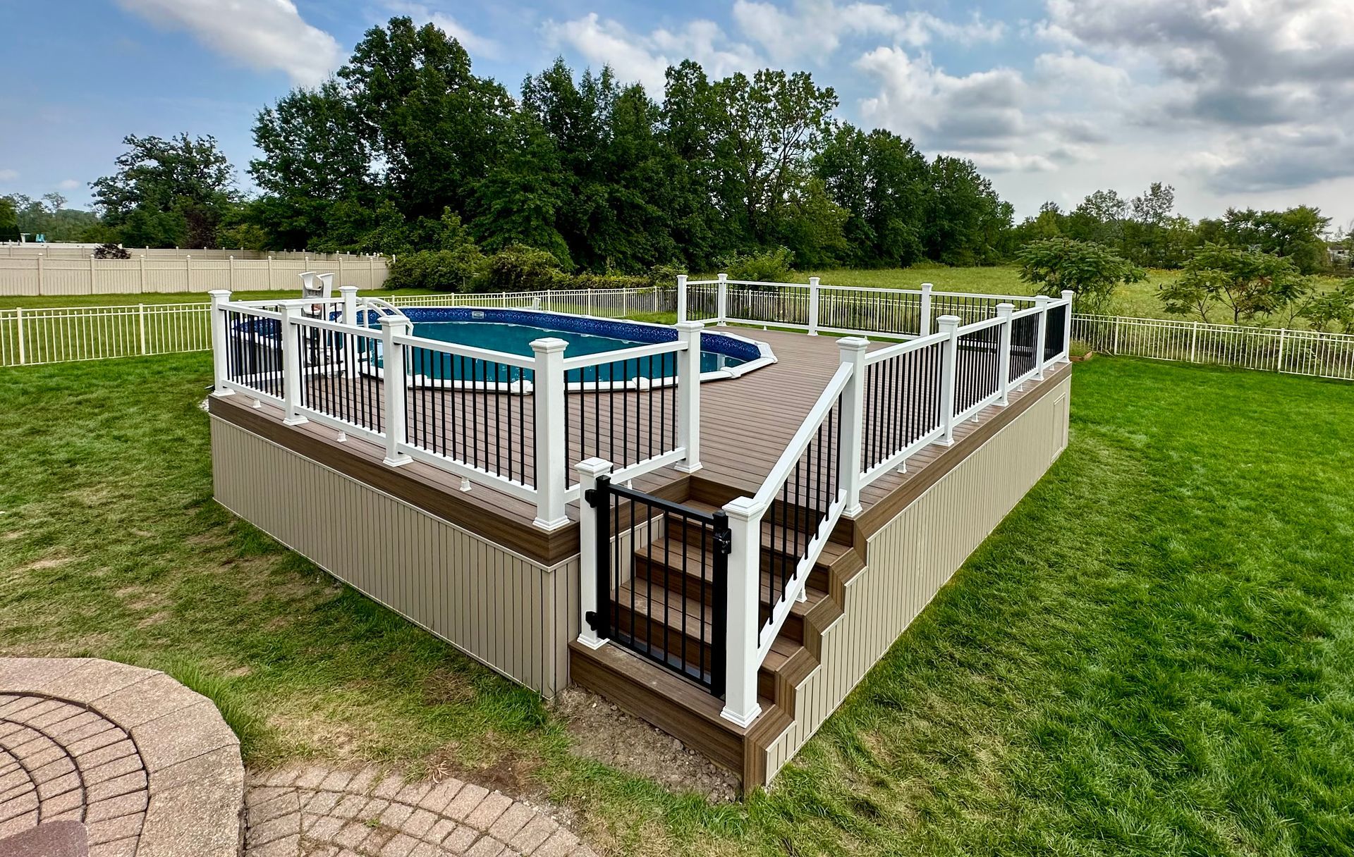 Above-ground pool on a deck with white railings and a small black gate. Green grass and trees in the background.