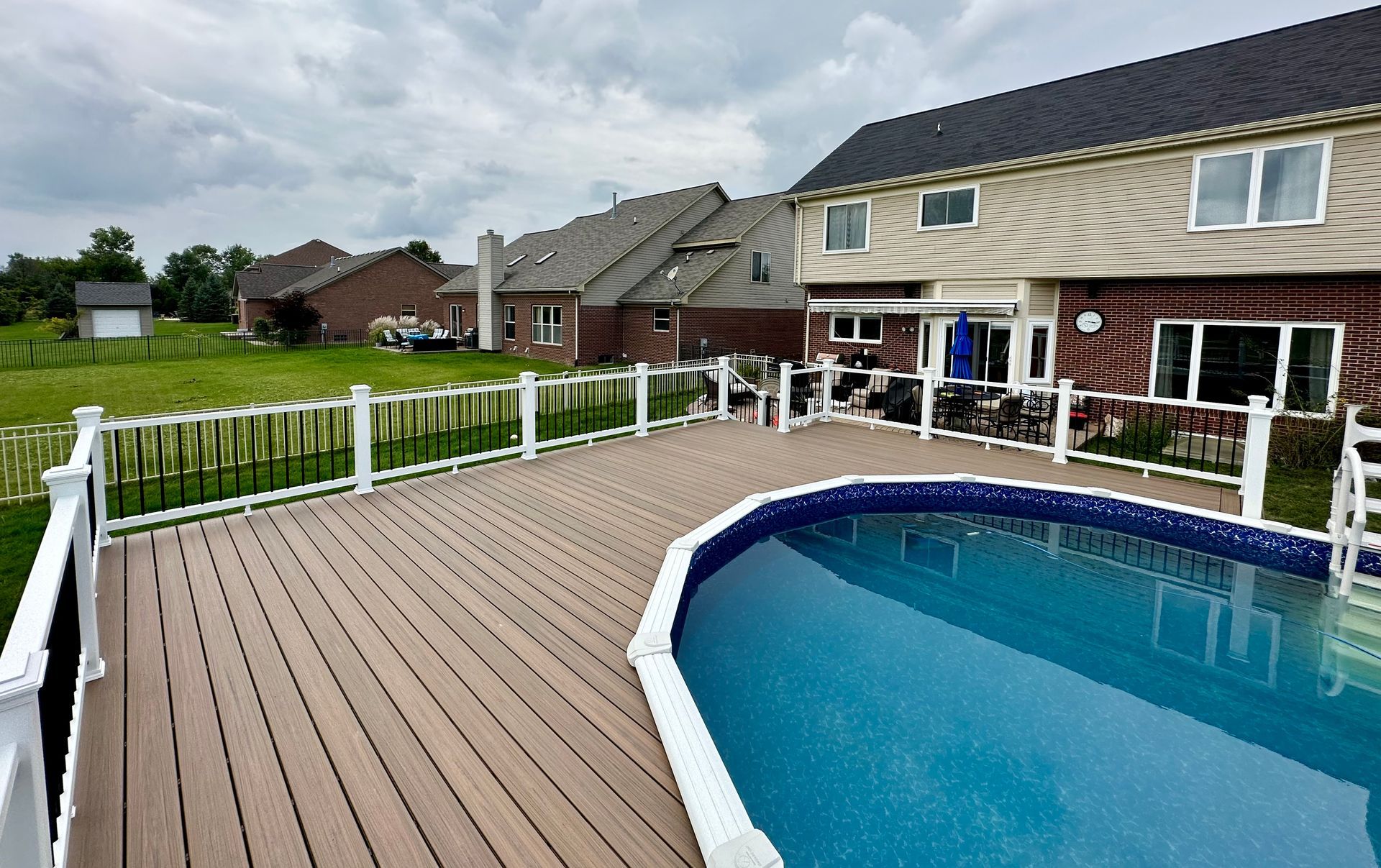 A backyard with a pool and deck. A two-story house is in the background on a cloudy day.