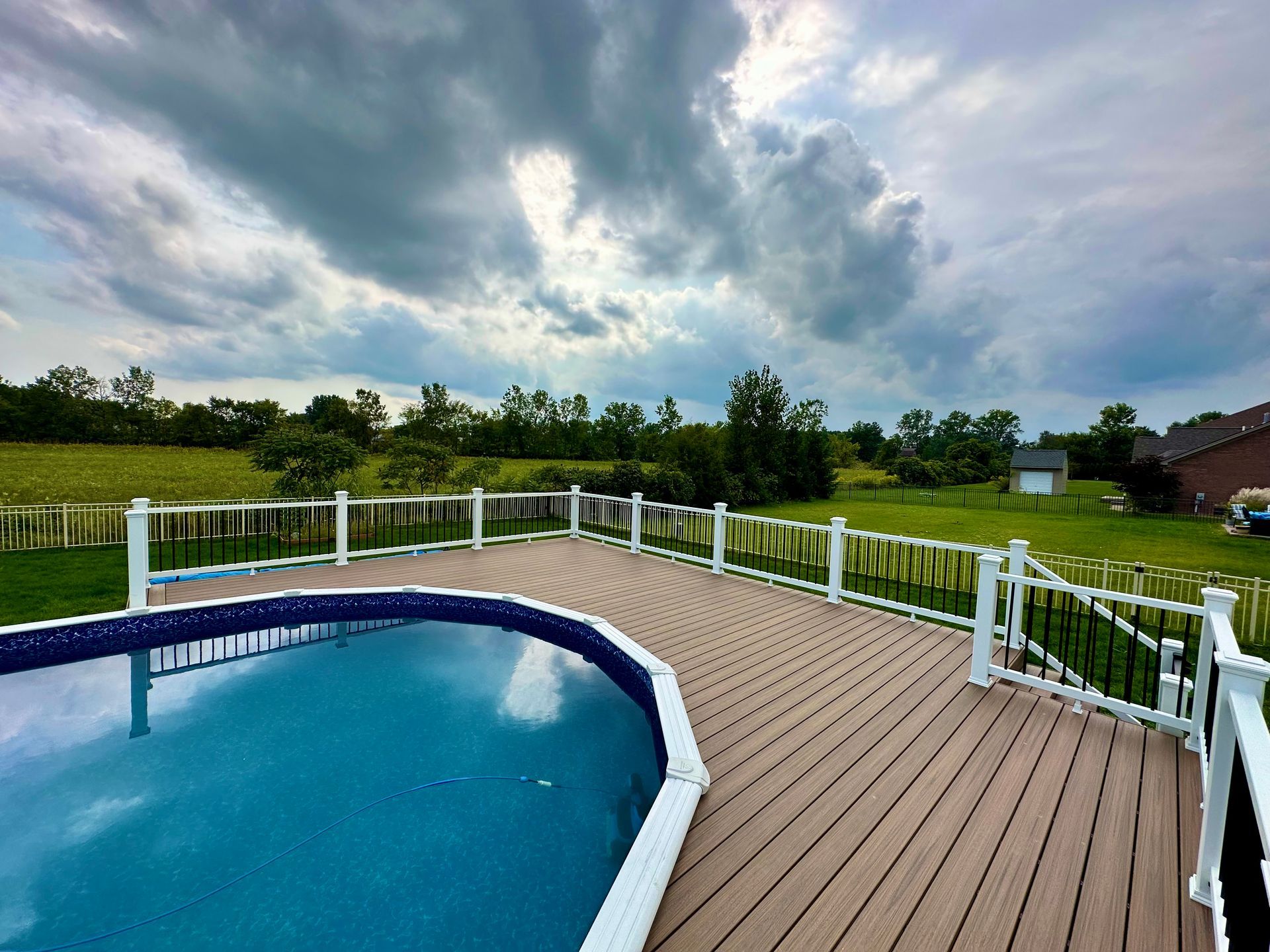Pool with brown deck and white railing, overlooking a green field under a cloudy sky.