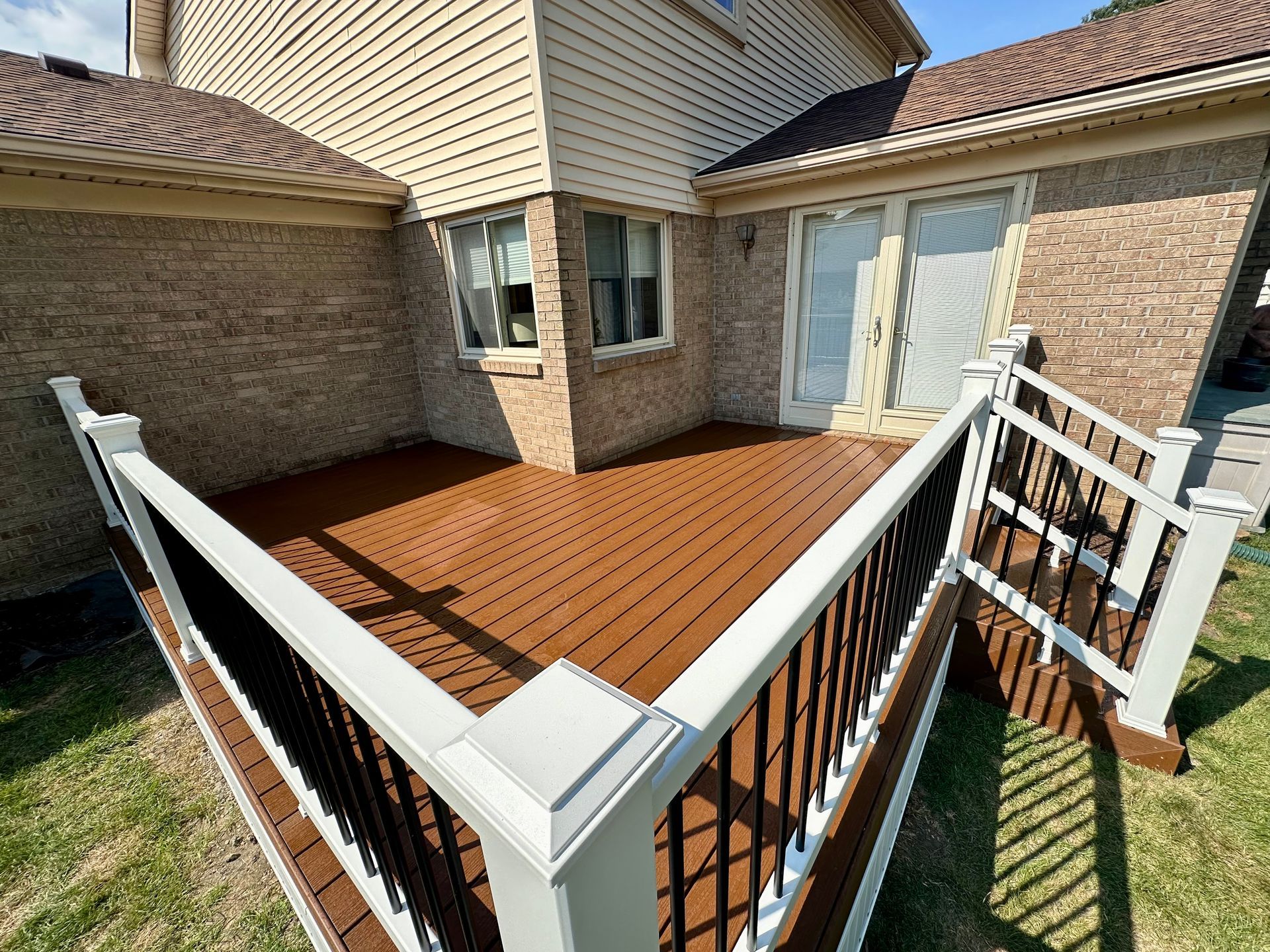 Brown deck with white and black railings, beside a brick house with glass doors.