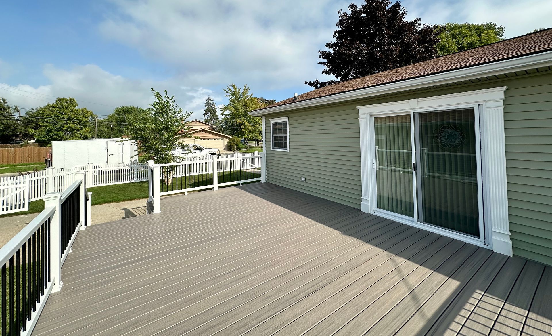 Gray deck with black railing, light green house with sliding glass doors, white fence, and green trees.