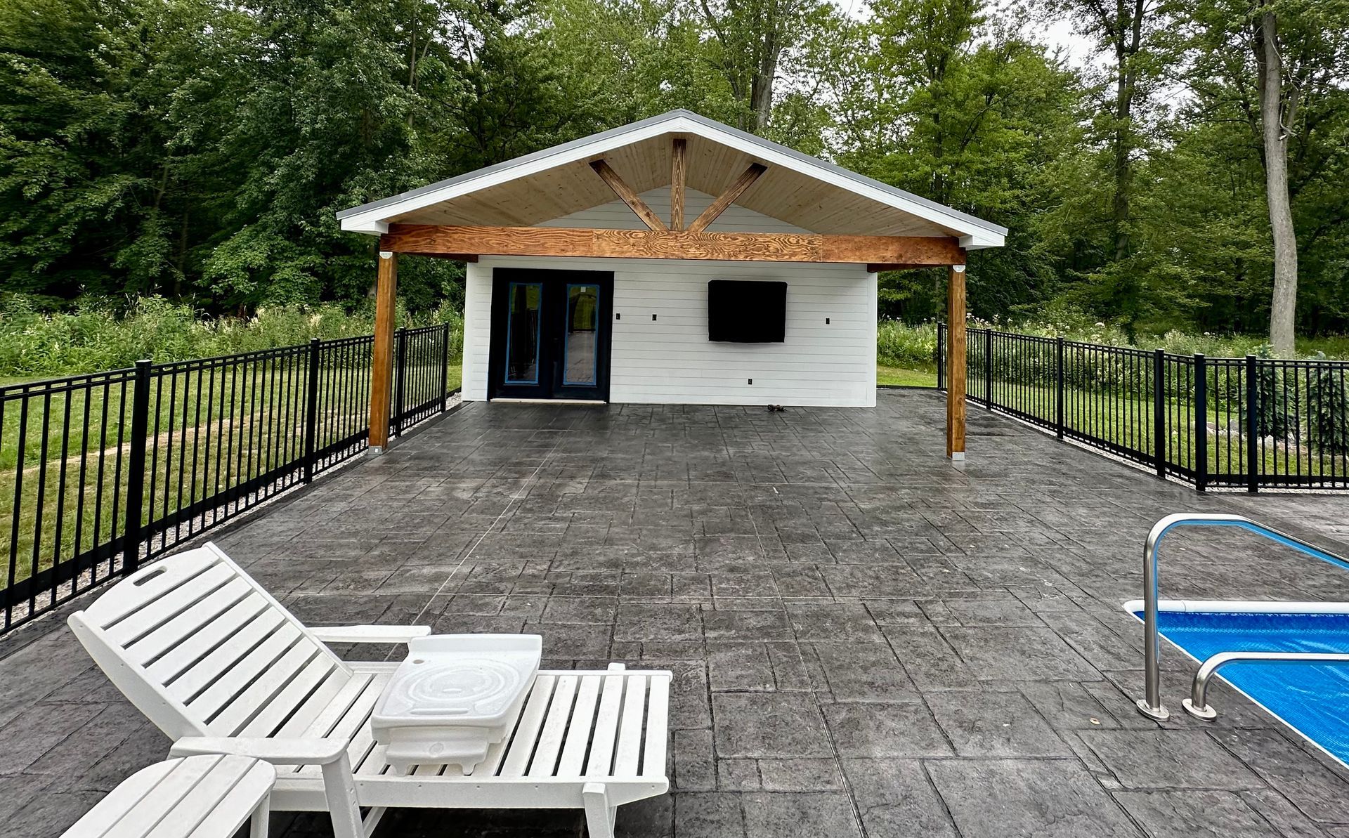 White building with a wooden overhang, on a gray deck next to a black fence and pool; white lounge chair.