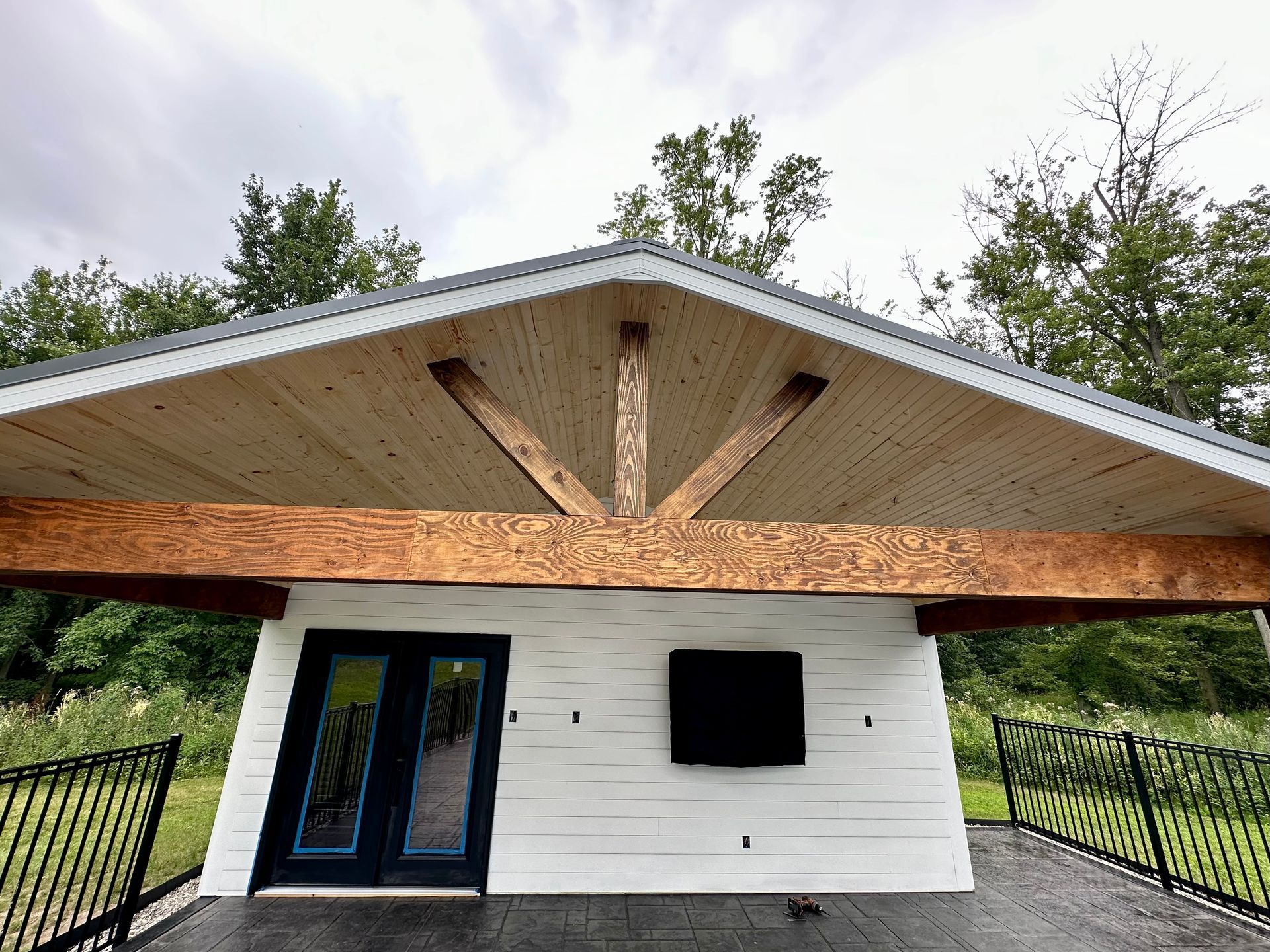 White building with wooden beams supporting a peaked roof. Black doors, a TV, and black railing. Green trees.
