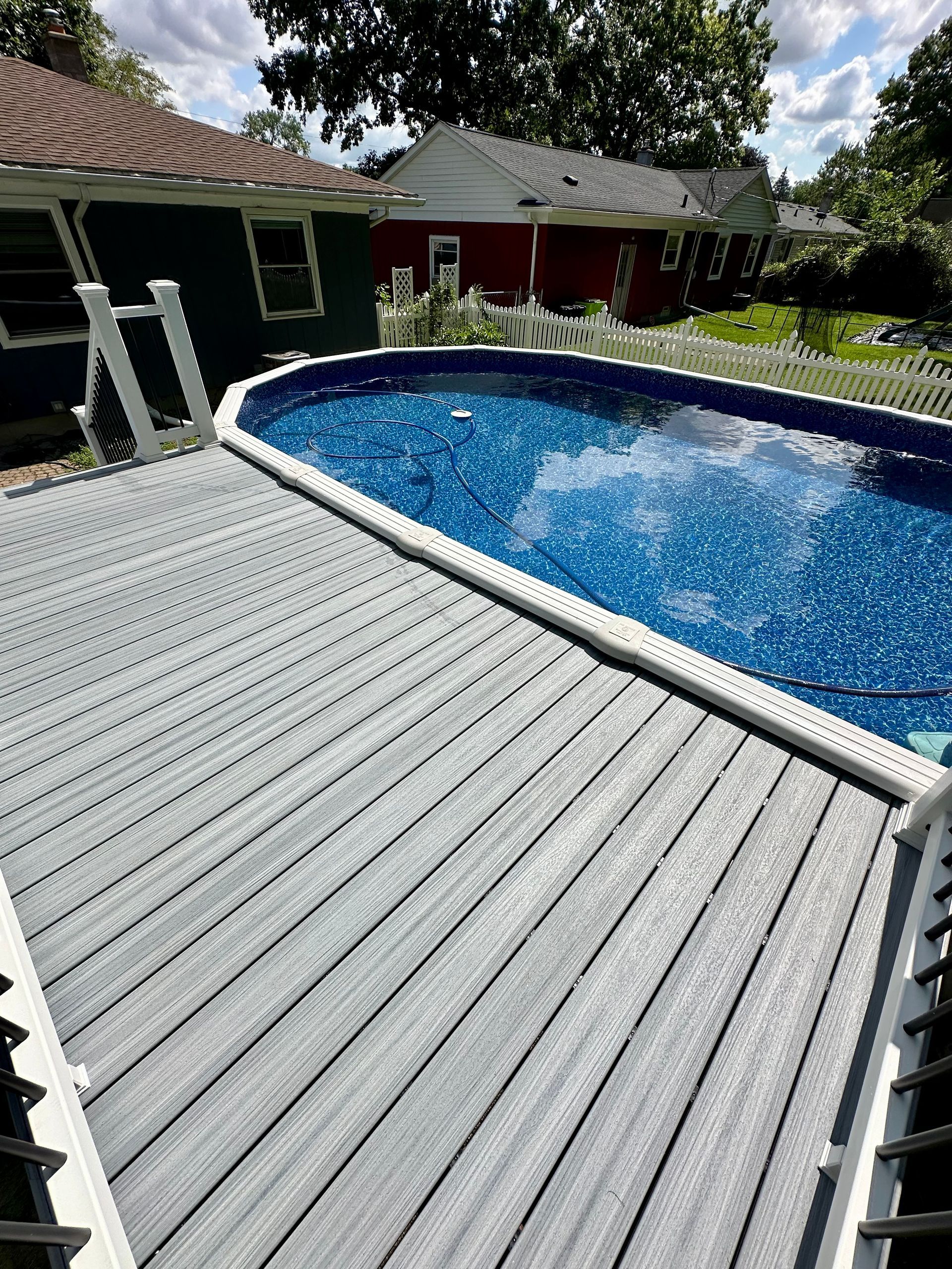 Above-ground pool with blue water and gray deck. Houses and trees are in the background.