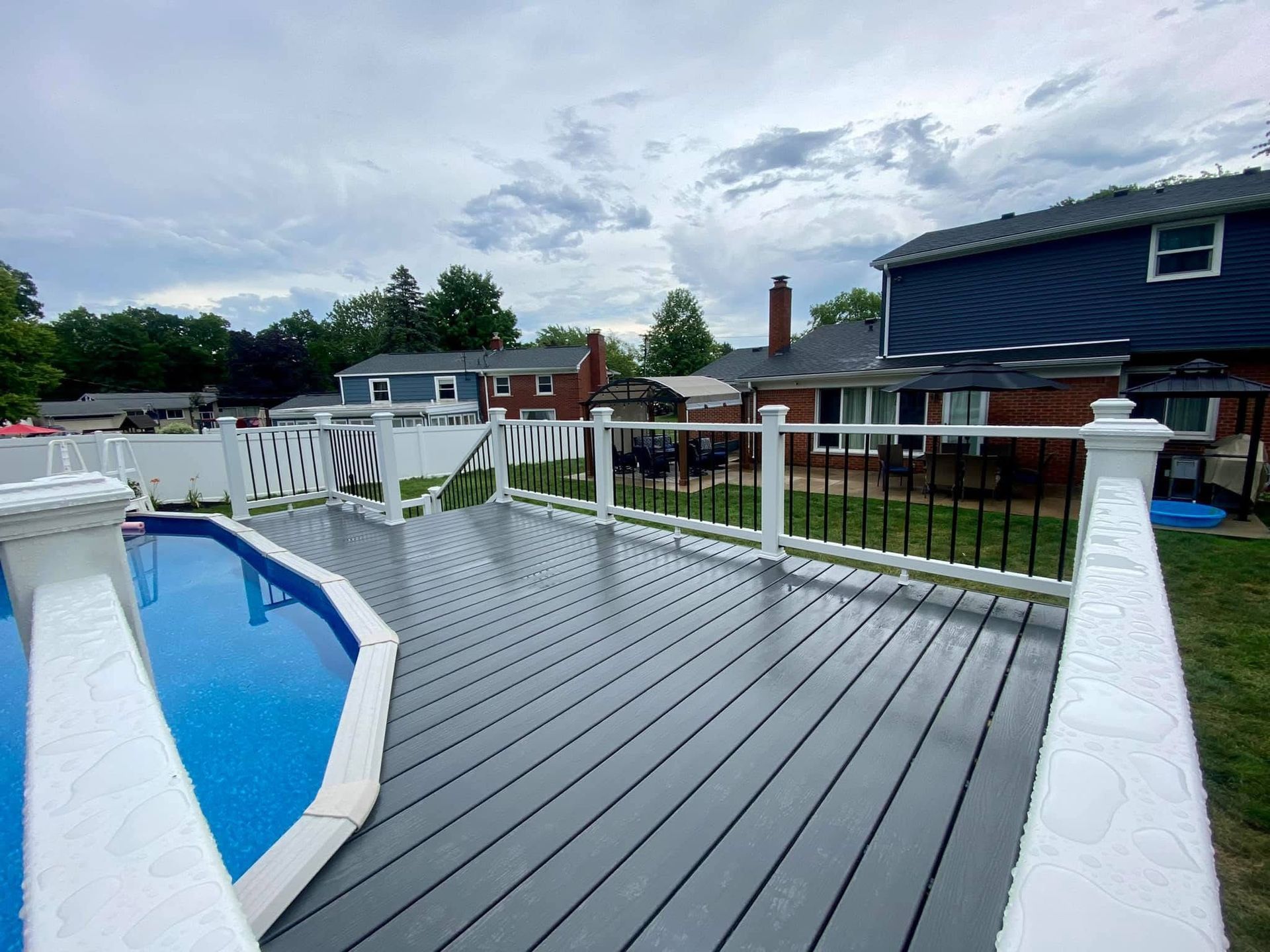 Gray composite deck with white railings surrounds a blue above-ground pool. Houses and cloudy sky in background.