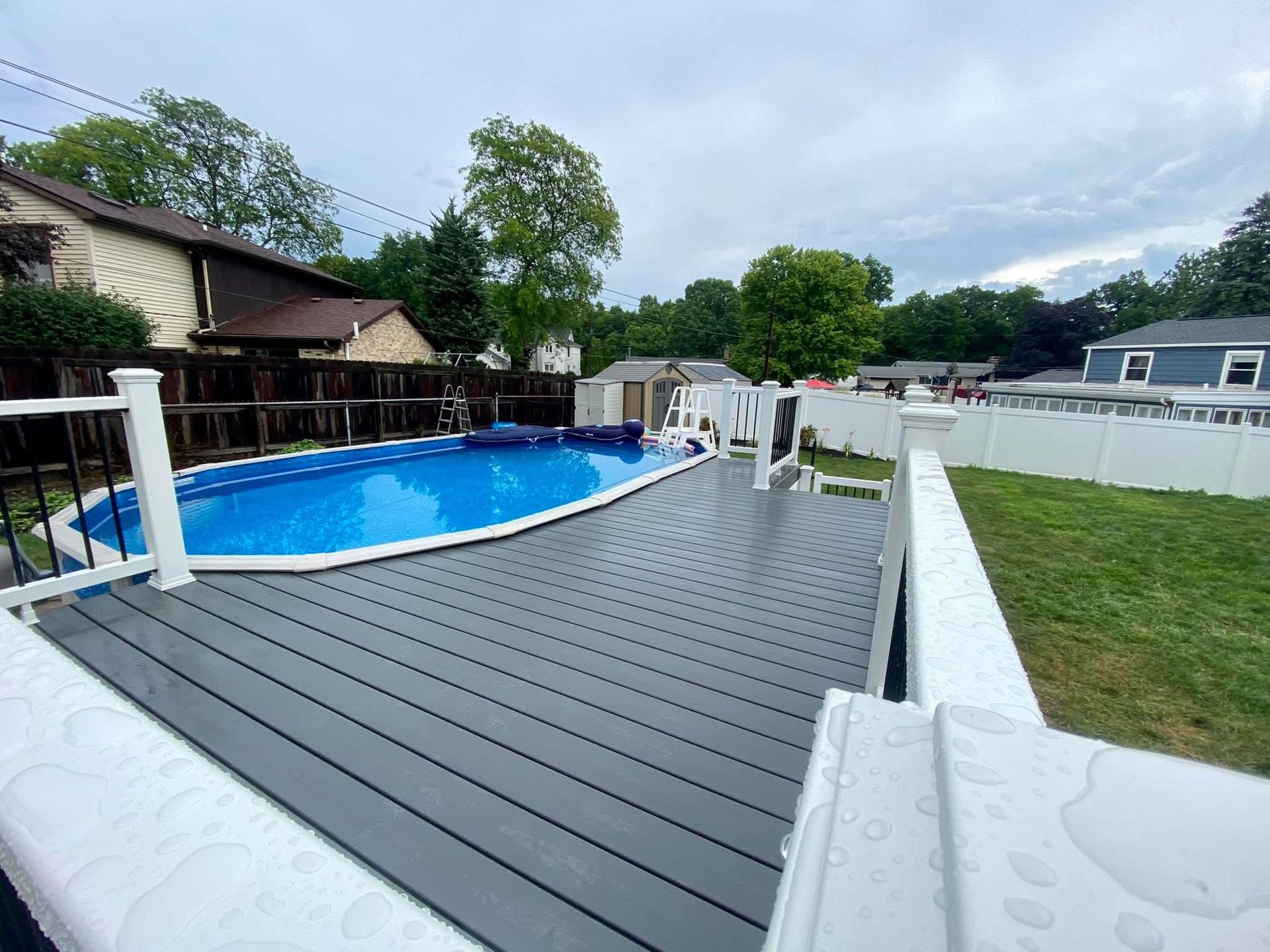 Backyard with above-ground pool and gray deck, white fence and railing, on a cloudy day.