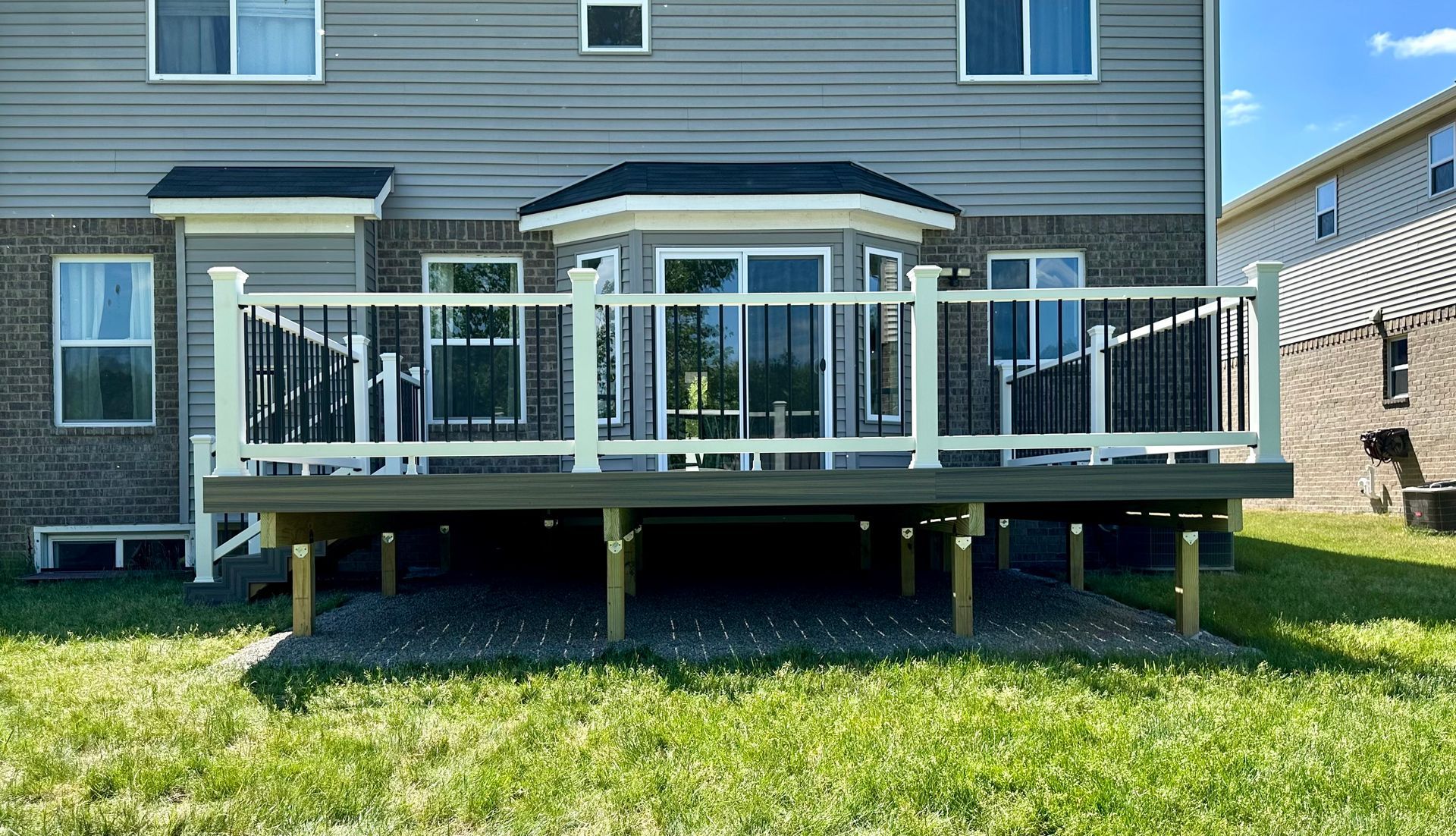 Backyard deck with white railing, black balusters, and gray decking, attached to a two-story house with a bay window.