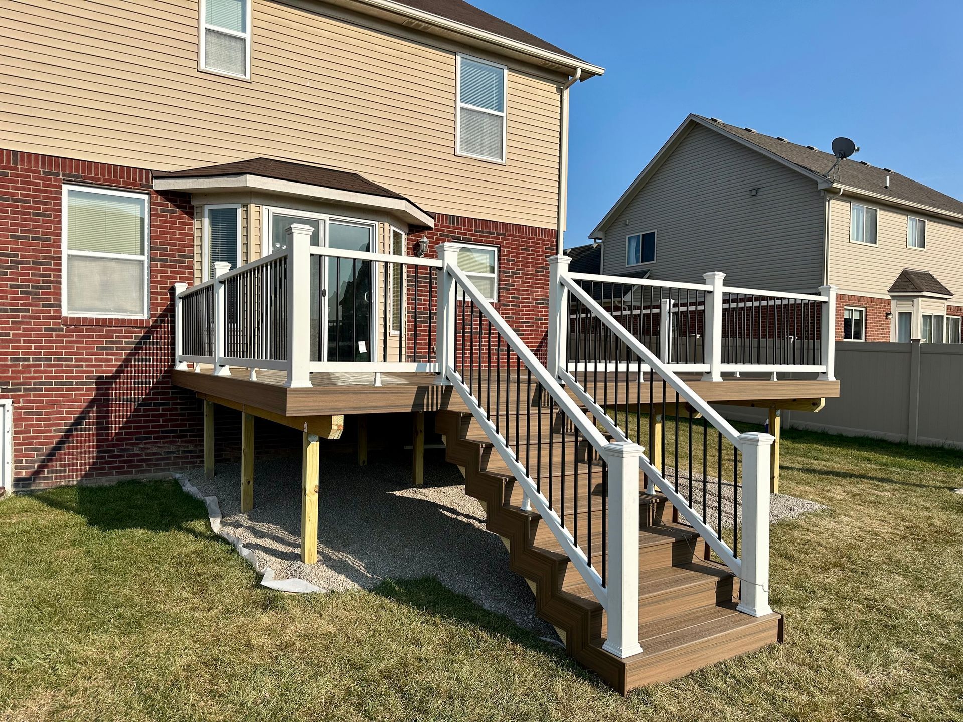 Backyard deck with stairs, built against a two-story brick and siding house. Brown deck, white railings, and black balusters.