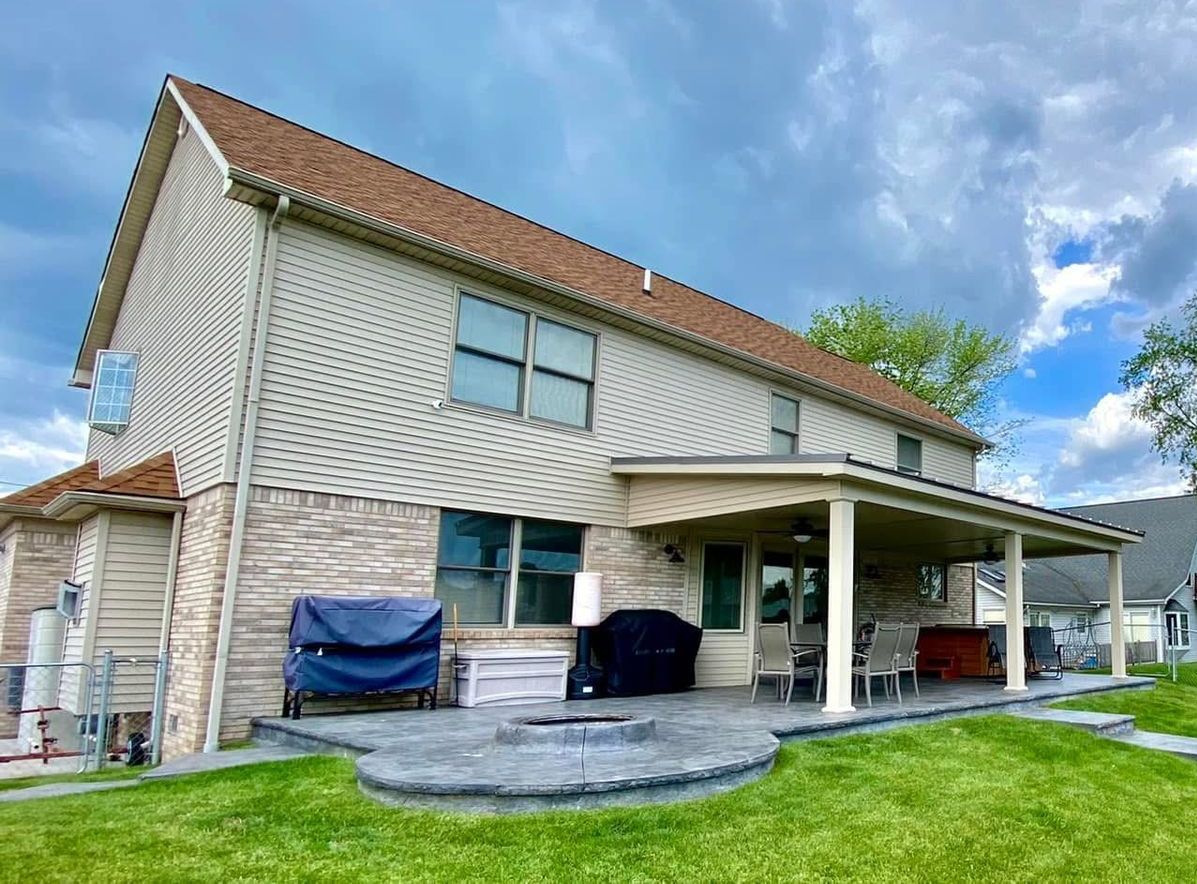 Two-story house with tan siding, brown roof, and covered patio with seating; cloudy sky overhead.