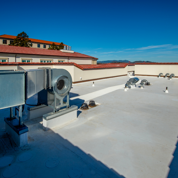 Rooftop with HVAC equipment, white membrane, against a sunny blue sky, building in background.
