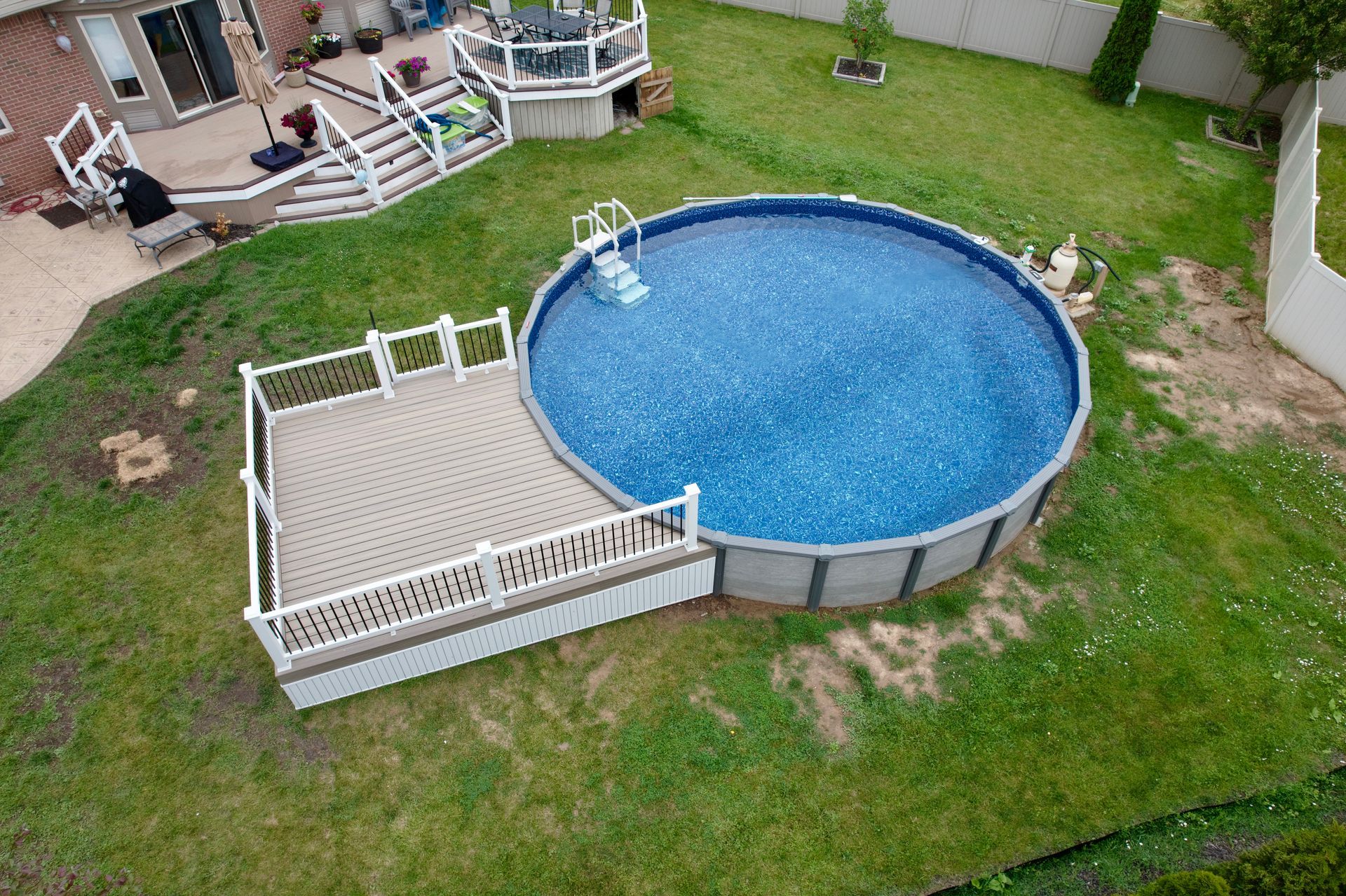 An above-ground pool with a deck in a backyard. The pool has blue water. Deck is made of composite wood with white railings.