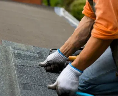 Roofer in orange shirt and gloves installing gray asphalt shingles on a roof.