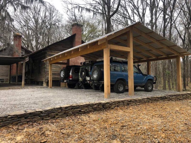 Two cars are parked under a wooden carport in front of a house.