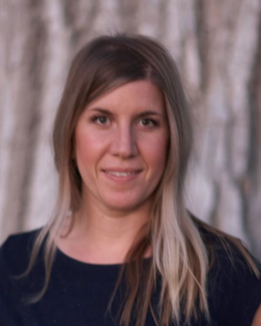 A woman in a blue shirt is smiling in front of a tree