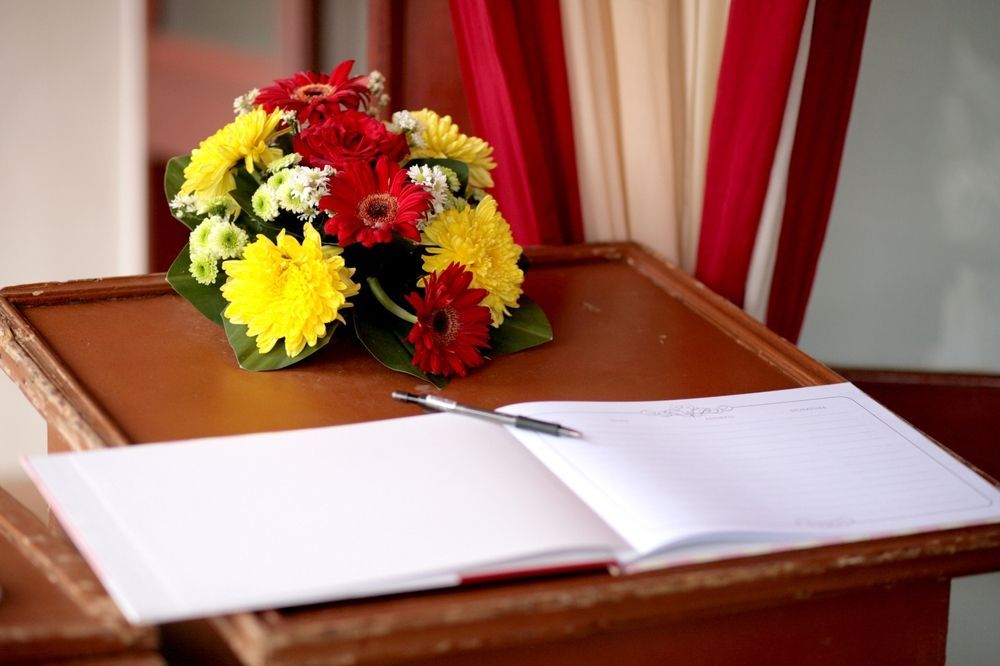 A bouquet of flowers sits on a wooden table next to an open book and a pen.