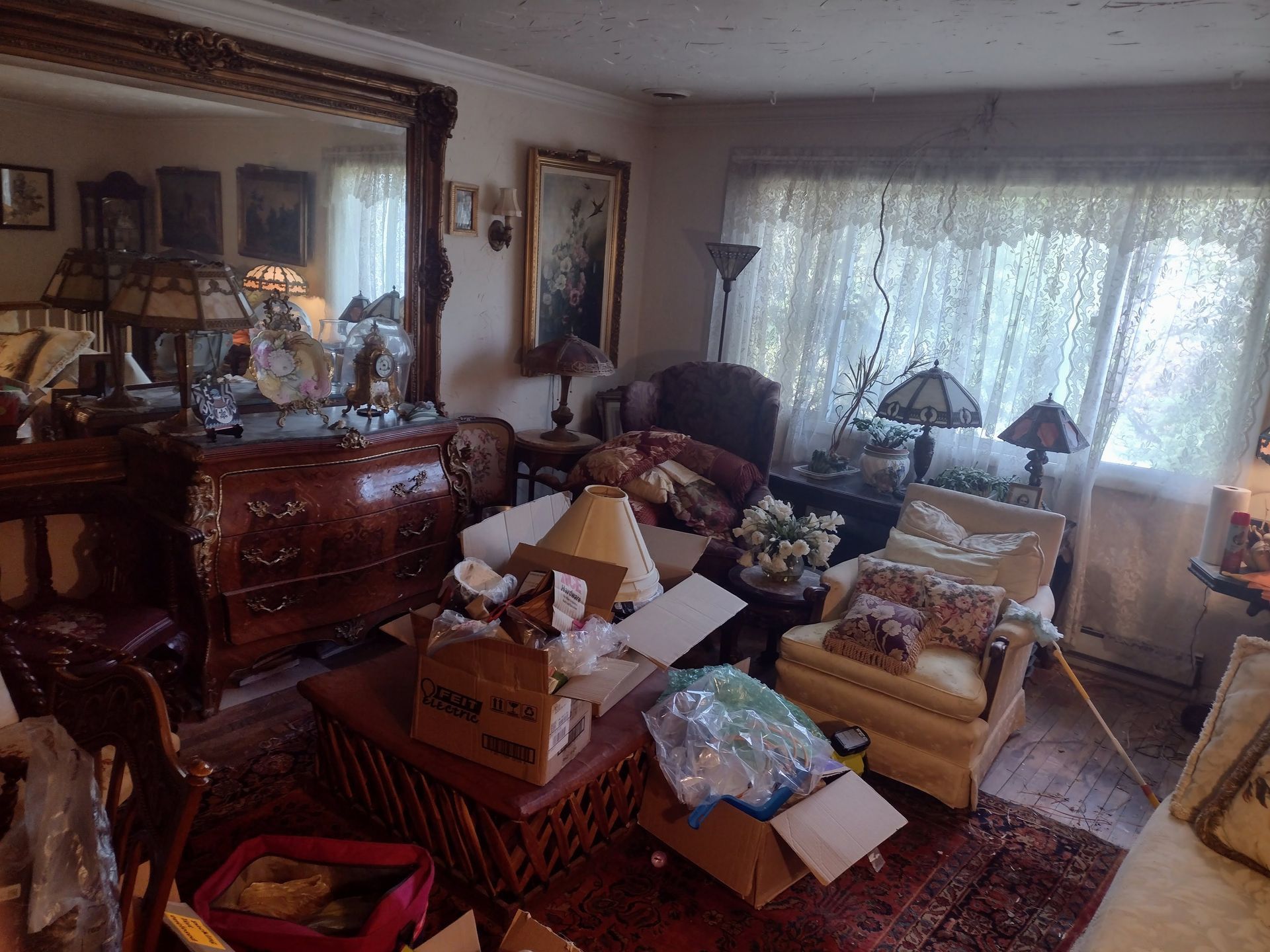 Cluttered living room with antique furniture, mirrors, lamps, and boxes. Red rug, lace curtains, and ornate decor.