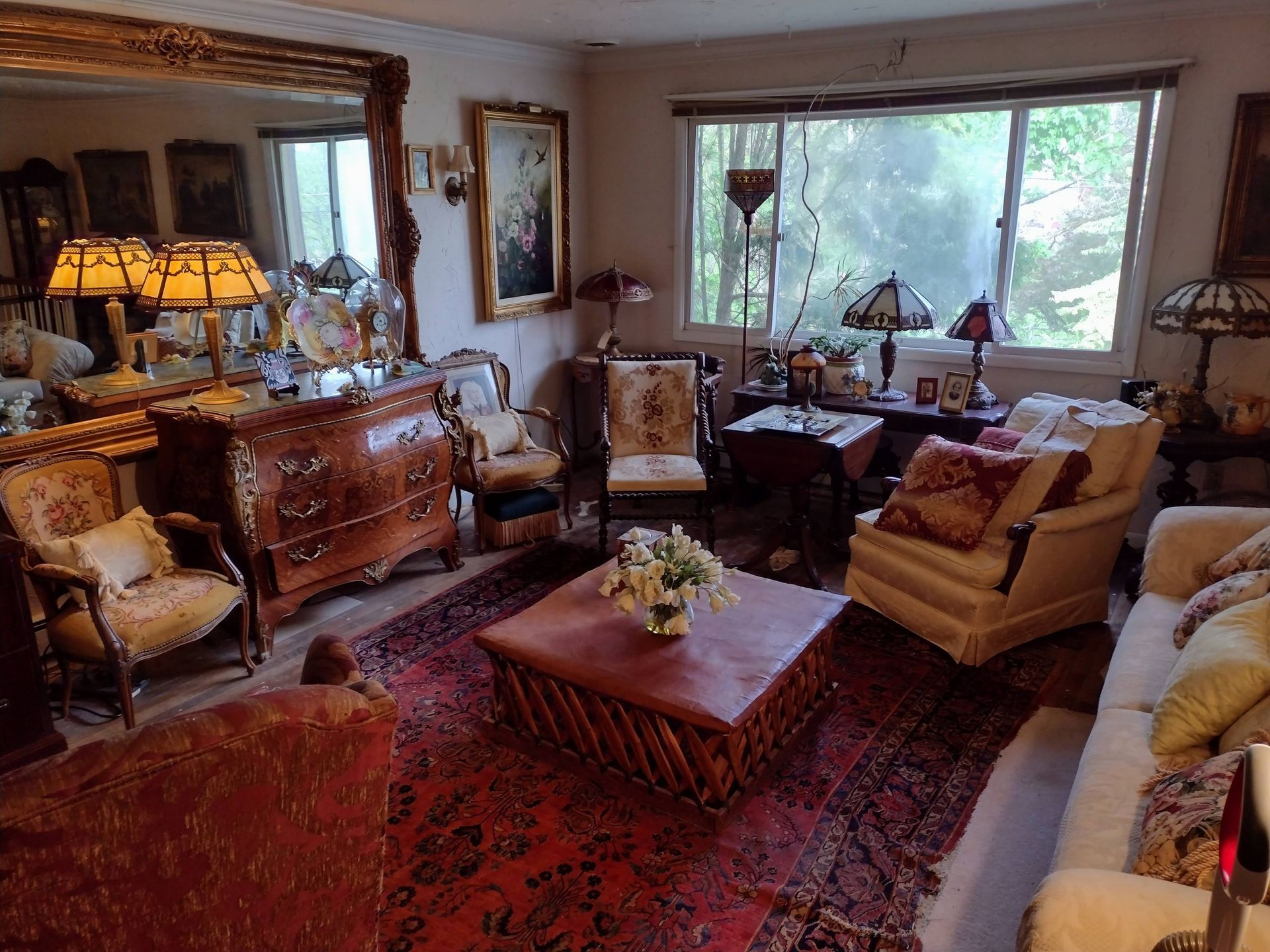A cluttered living room with antique furniture, oriental rug, ornate lamps, and large mirror.