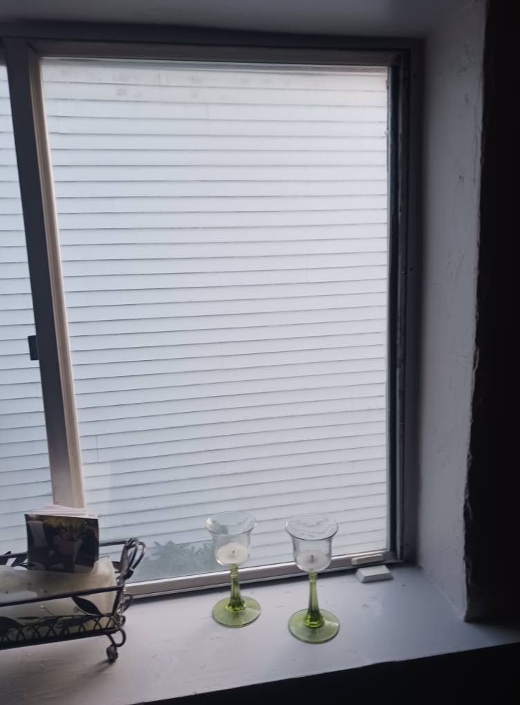 Two green-stemmed glass candle holders on a windowsill. Outside, a white-sided building is visible through the window.