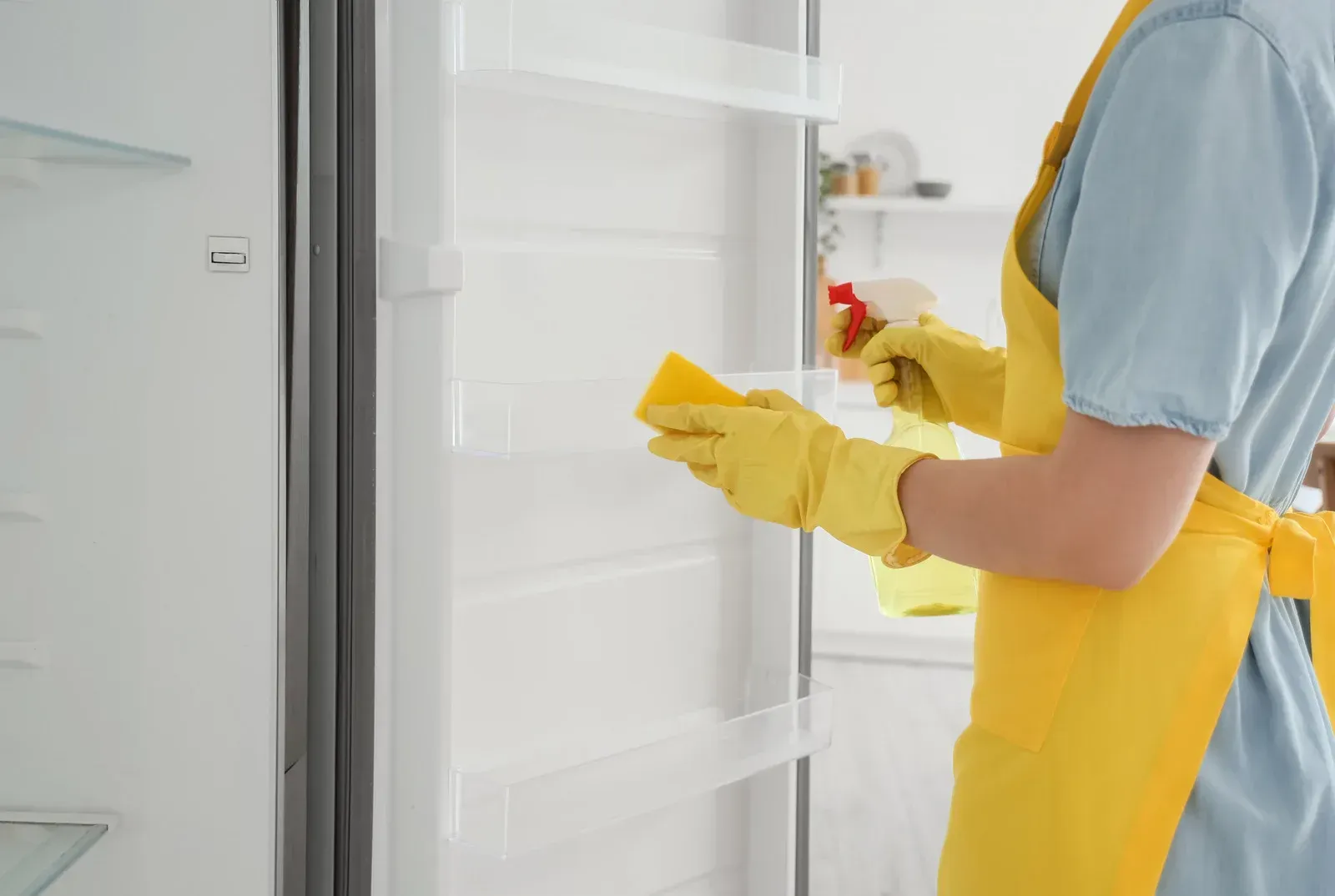 Woman in gloves cleaning an empty refrigerator with a sponge and spray bottle.