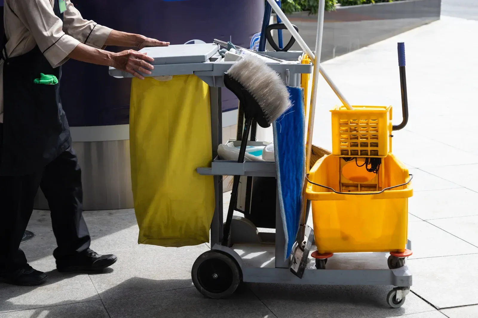 Person pushing a cleaning cart on a sidewalk. Yellow bucket, trash bag, and cleaning supplies visible.