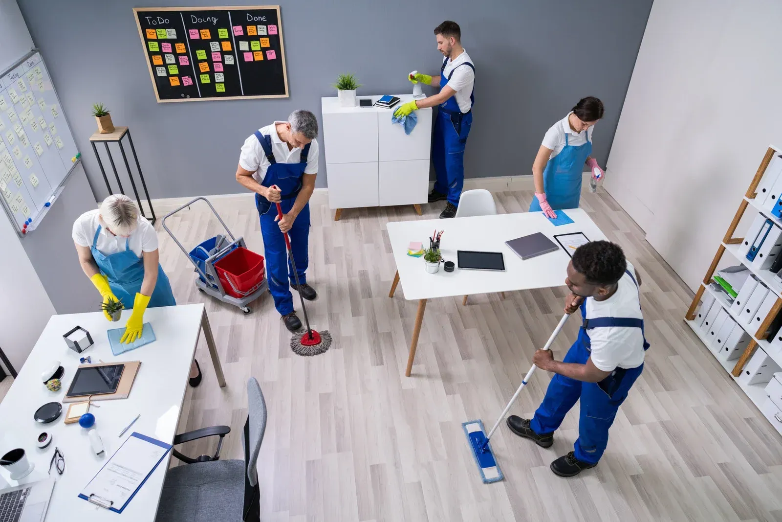 Office cleaners in blue uniforms clean desks, mop floors, and dust in an office setting.