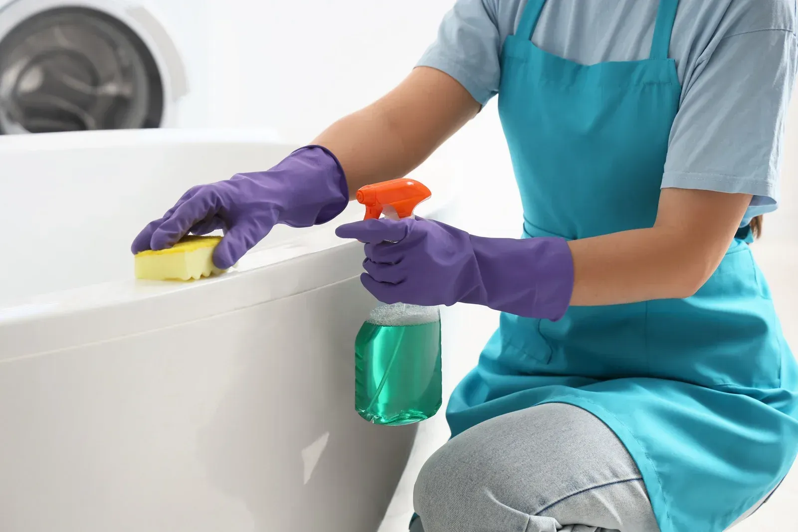 Person in purple gloves and teal apron cleans a white bathtub with a sponge and spray bottle.