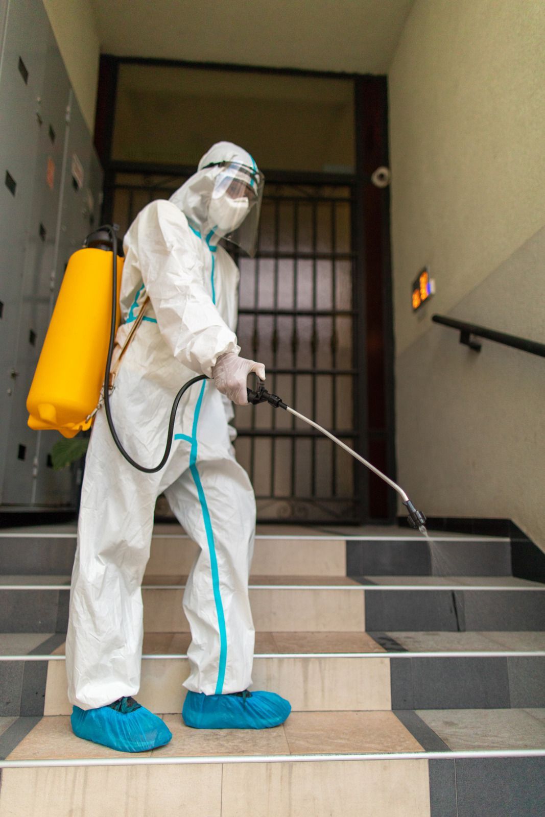 Person in hazmat suit sanitizing stairs with spray. Yellow tank, blue booties, and face shield.