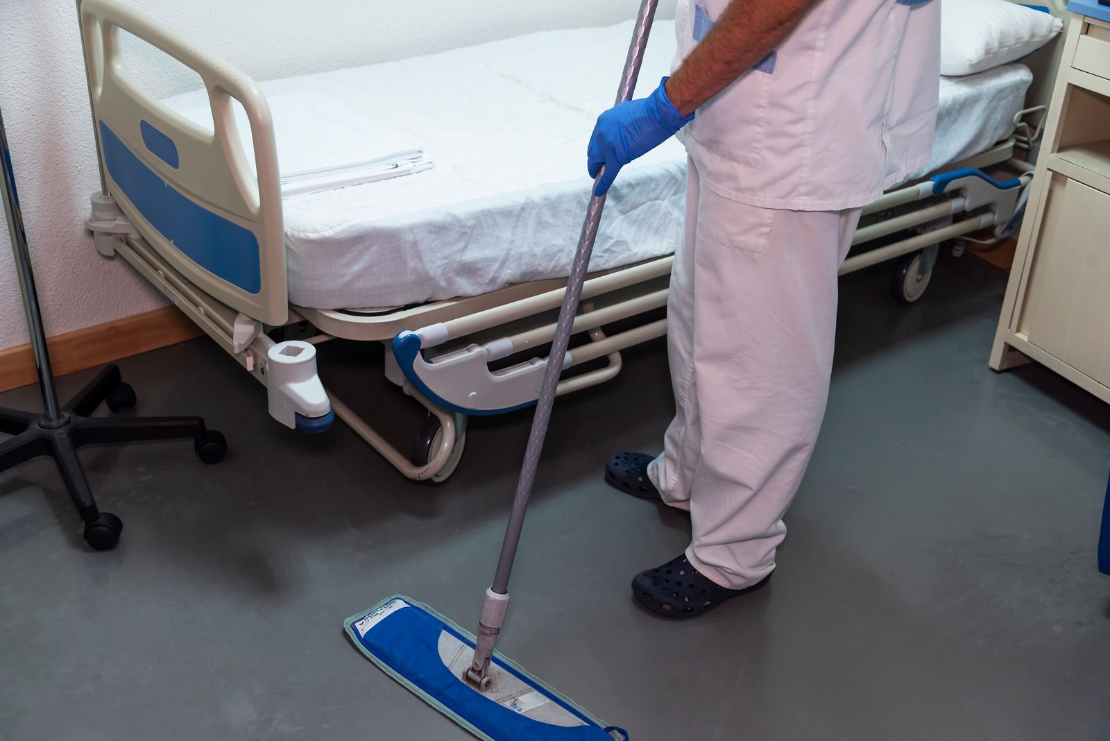 Person in scrubs mopping hospital floor near an adjustable bed.
